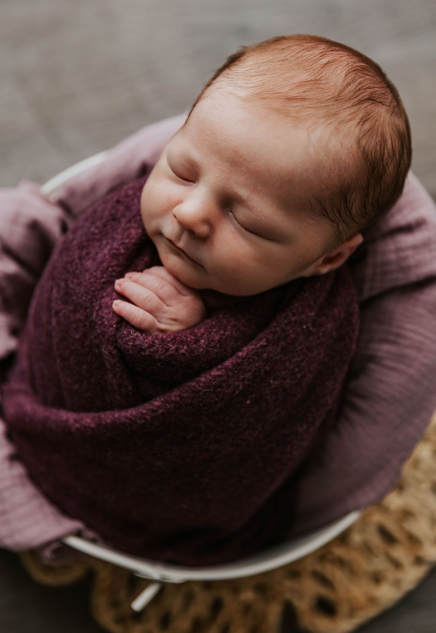 Sleeping newborn swaddled in plush purple fabric resting on a textured surface.