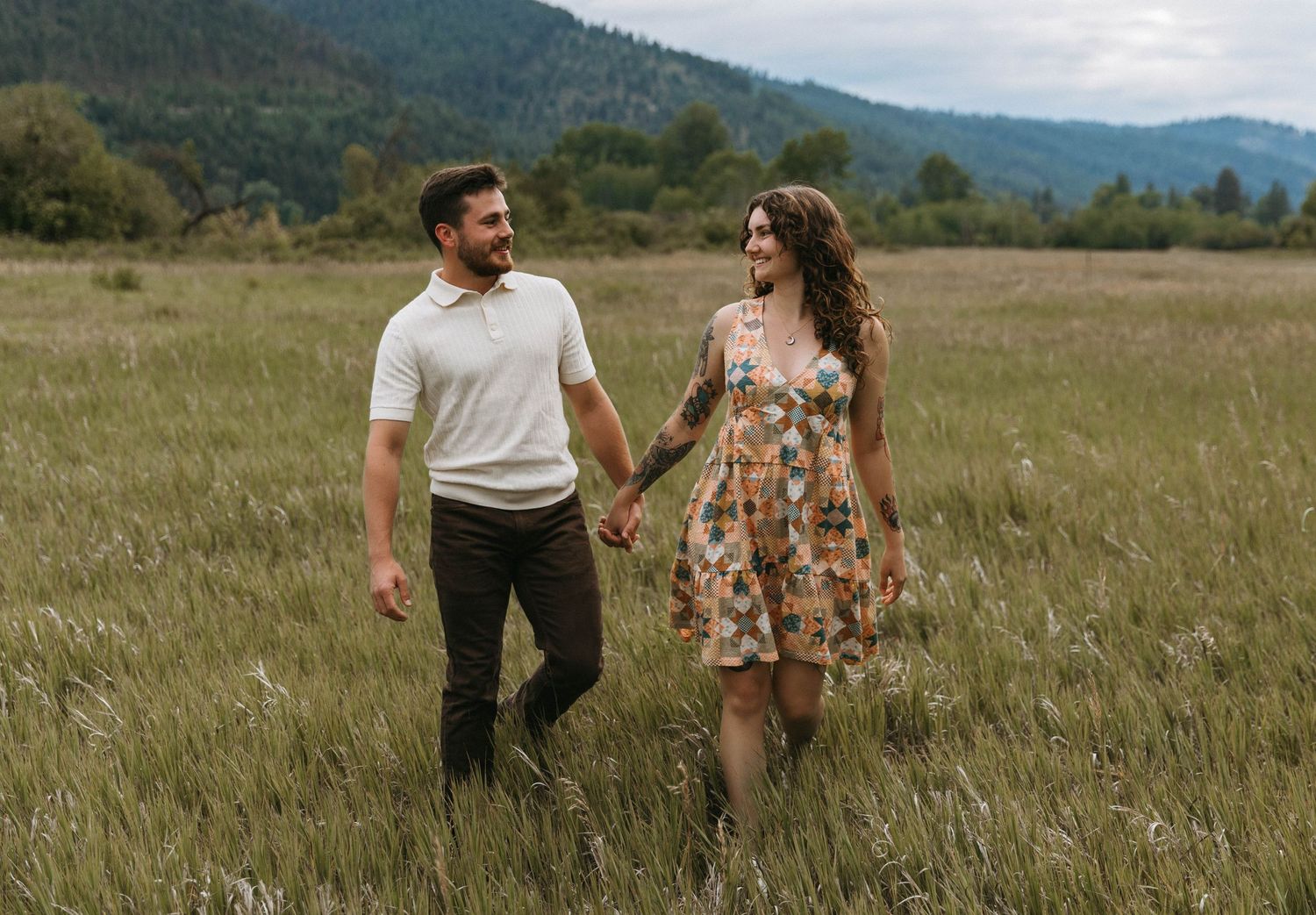 A couple holds hands while walking through a golden meadow with mountains in the background on a cloudy day.