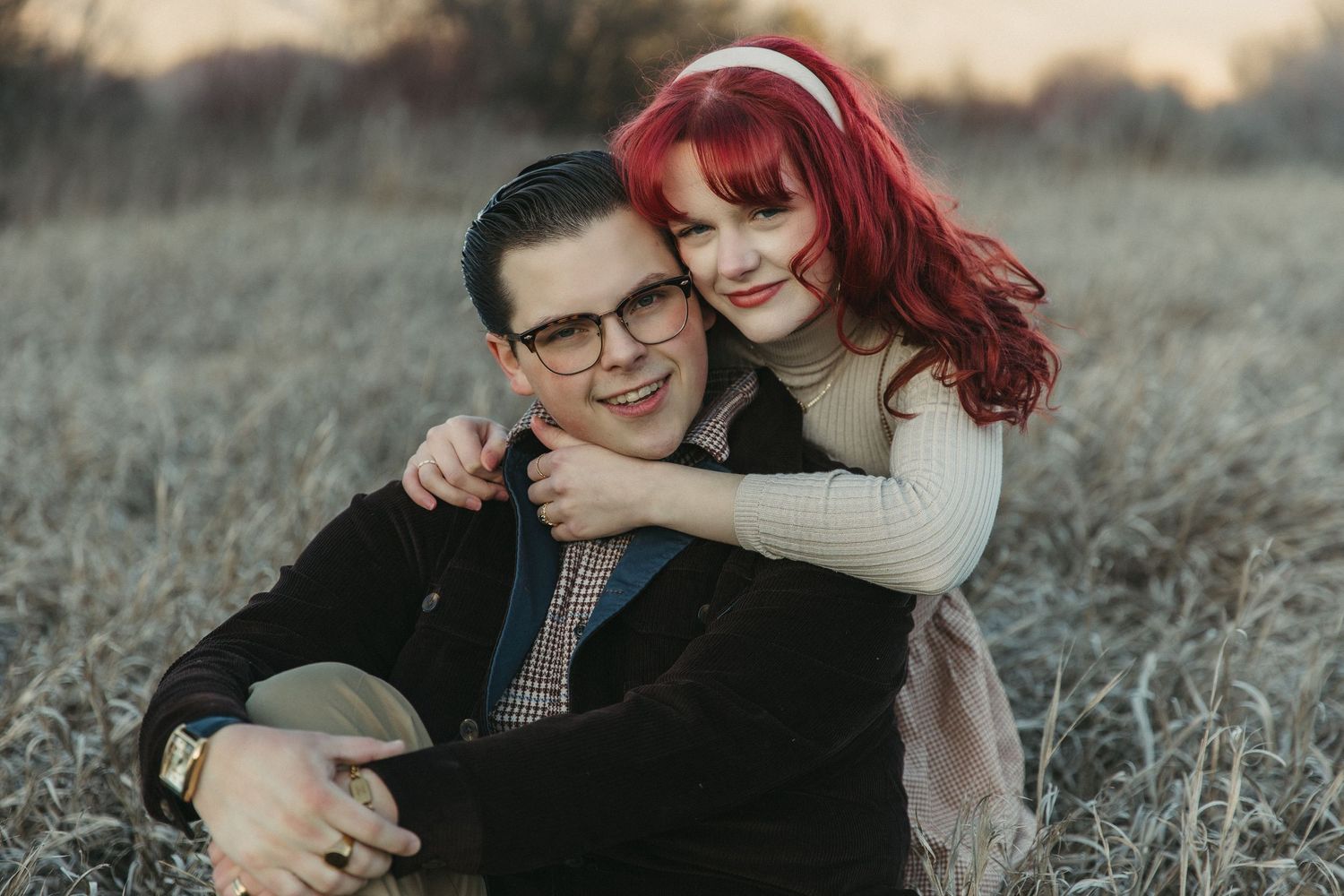 A man and red-haired girl smile warmly while sitting in a field, sharing a loving embrace during golden hour.