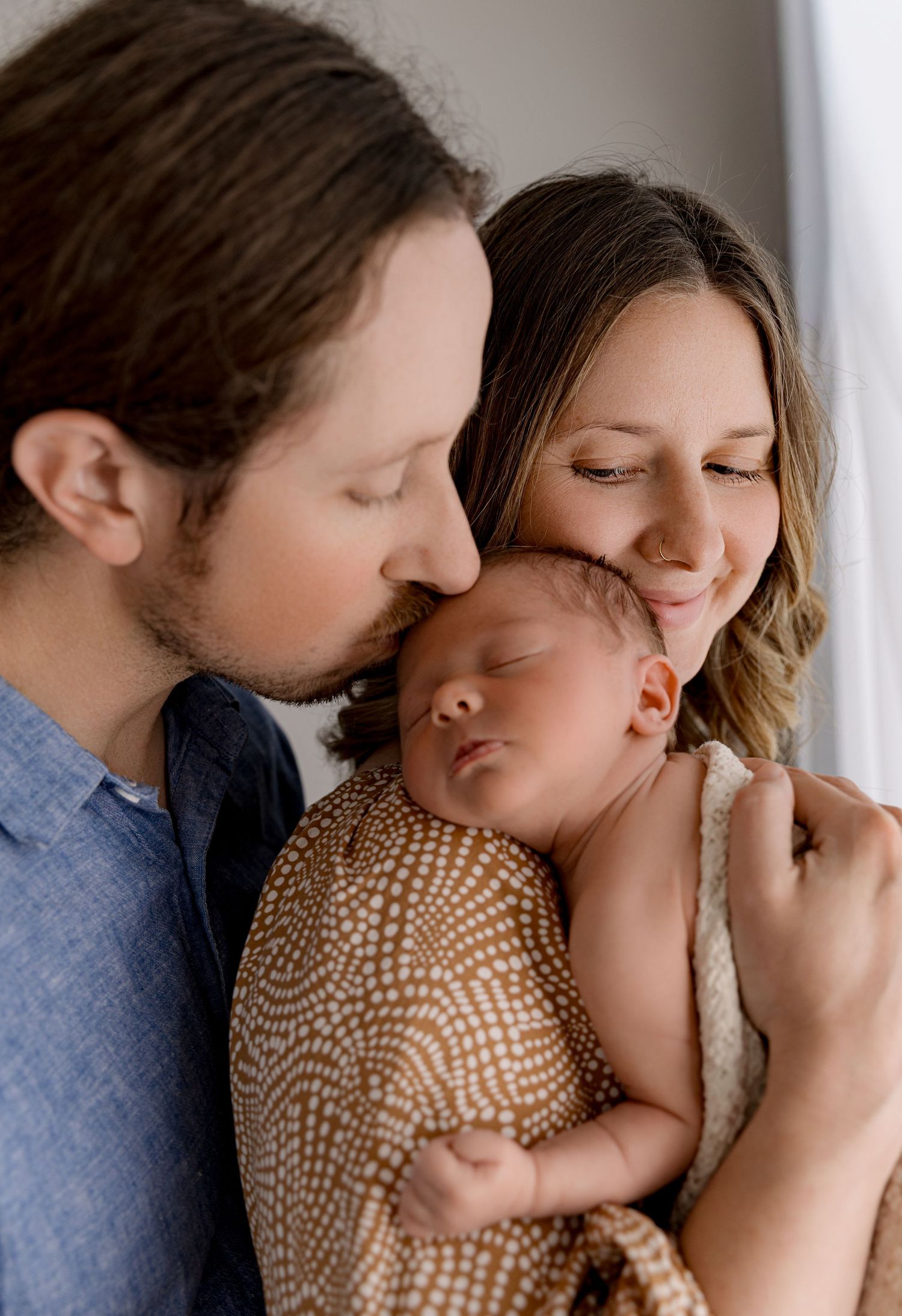 A couple tenderly kisses their sleeping newborn baby wrapped in a brown patterned swaddle blanket.