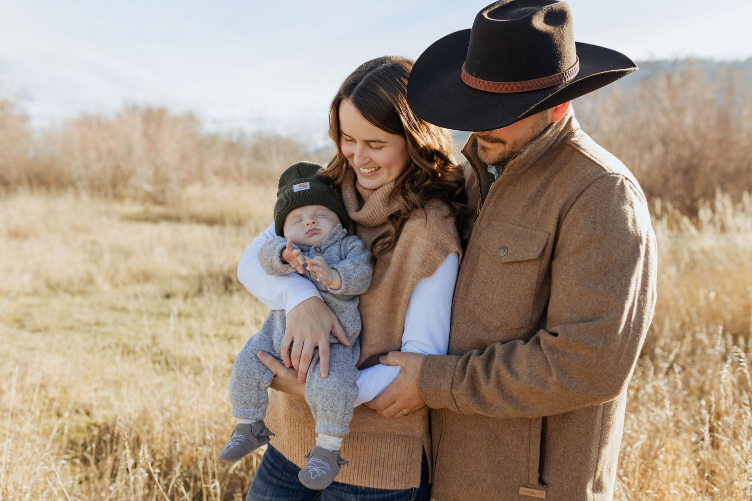A couple and infant stand together in golden grass fields with mountains in background wearing western attire.