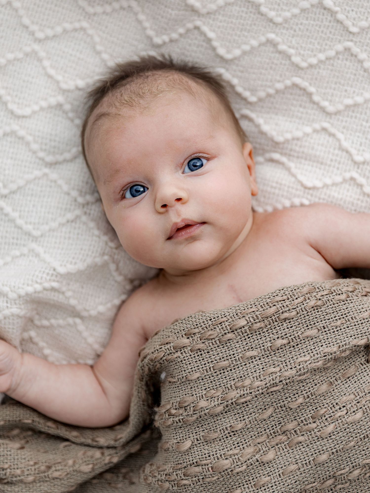 A baby lying on a textured white blanket, partially covered with a beige knit wrap, gazing up with bright blue eyes.