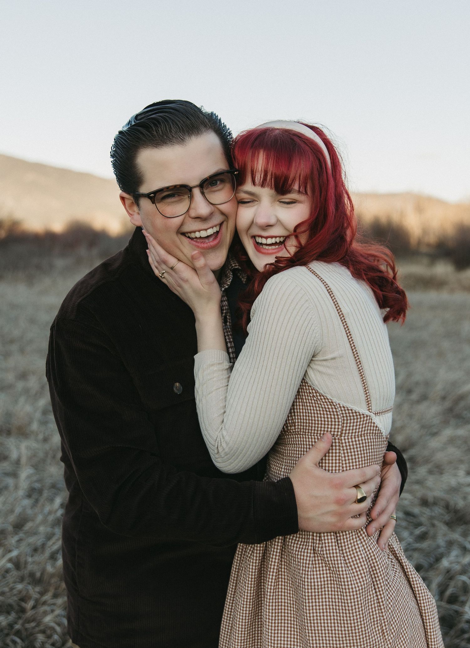 A couple embraces and laughs together in a desert landscape during sunset, wearing casual winter attire.