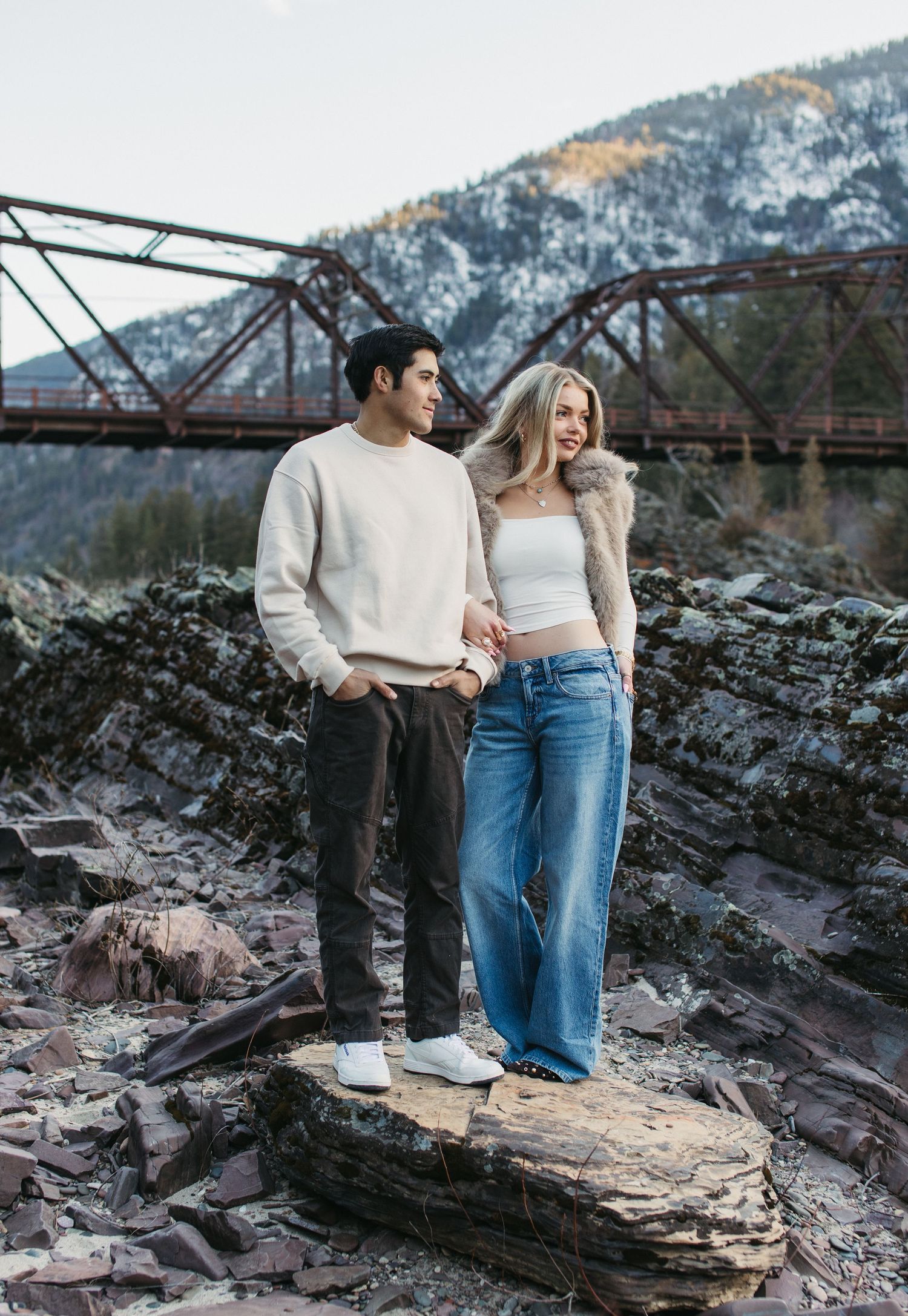A couple stands together on rocky terrain near a rustic bridge with snow-capped mountains in the background.