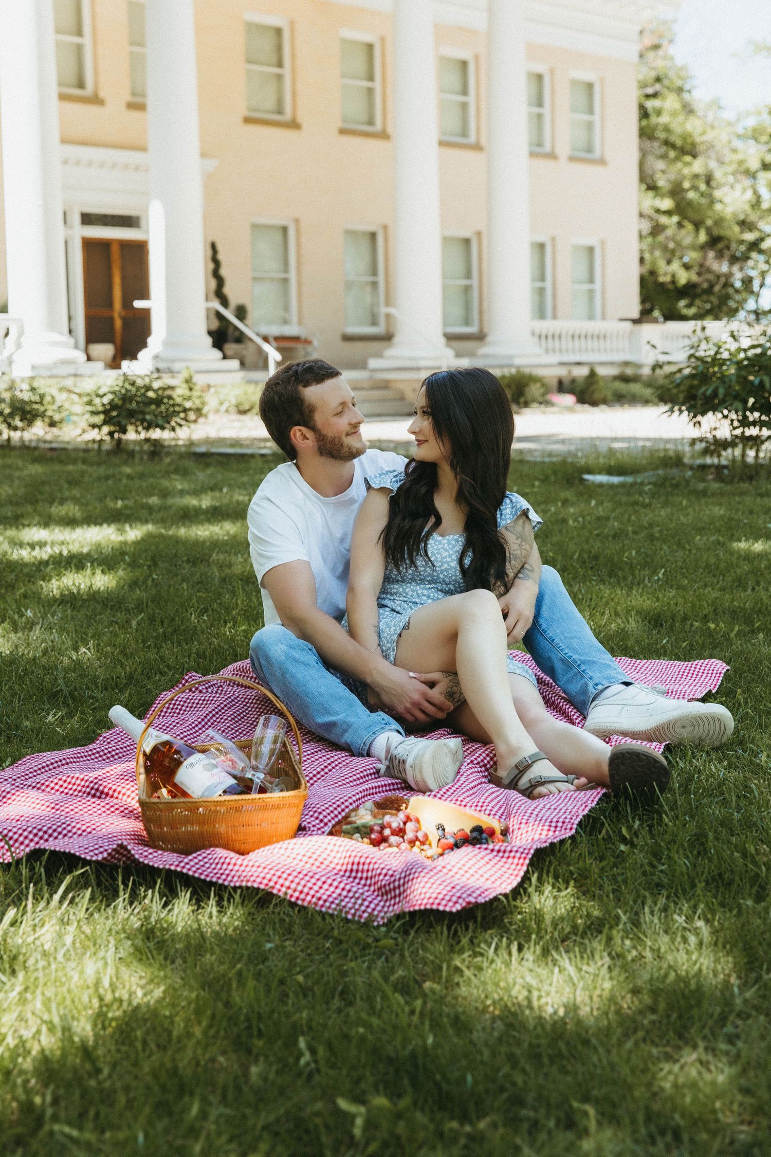 A couple relaxes on a red checkered picnic blanket while enjoying refreshments in the shade of a stately building.
