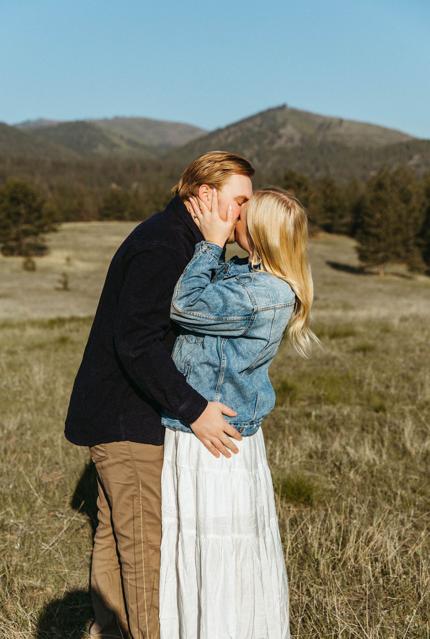 A couple embraces in a field with mountains in the background on a sunny day.
