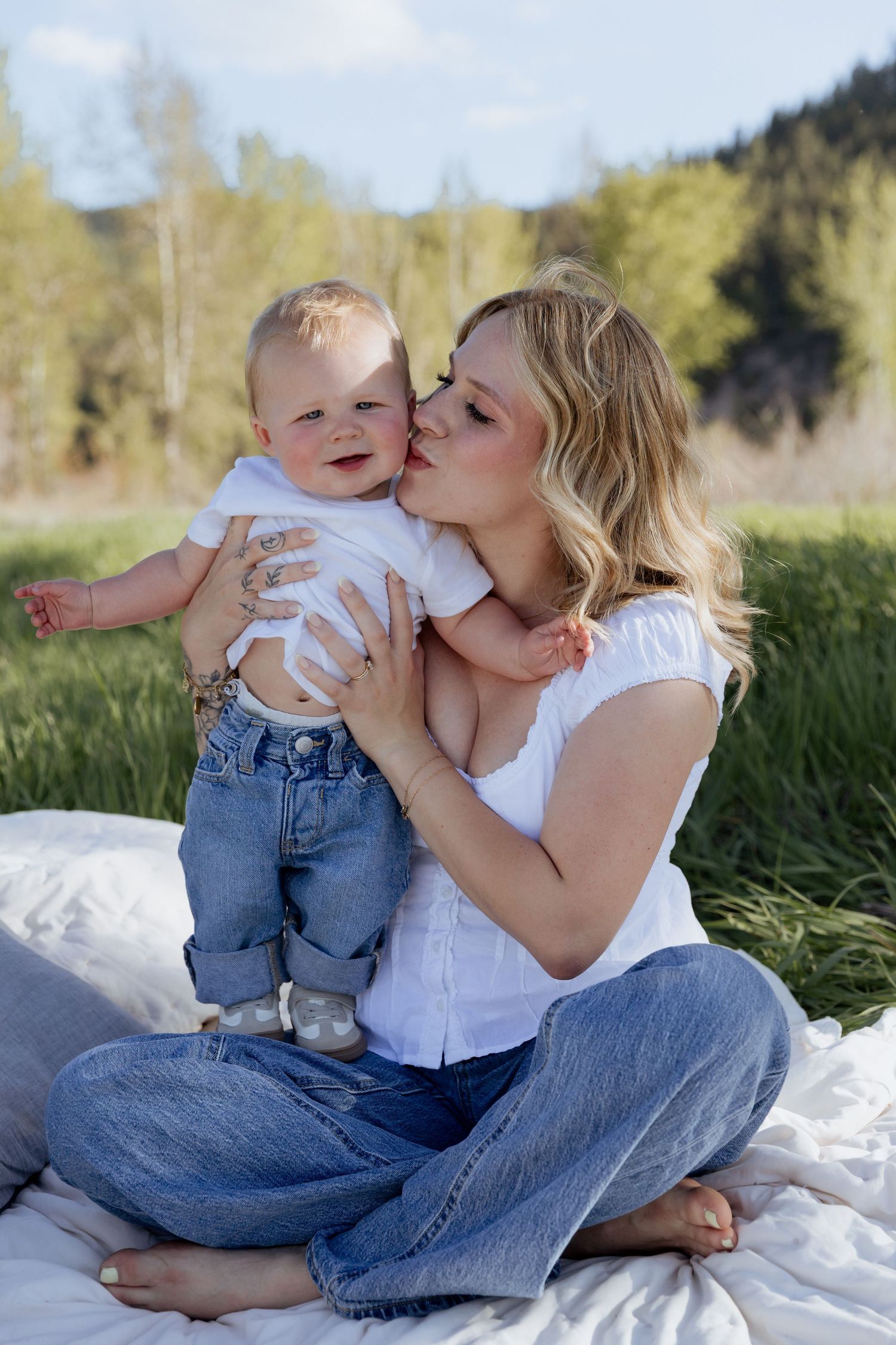 A blonde mother kisses her smiling baby dressed in jeans on a white blanket in a green meadow.