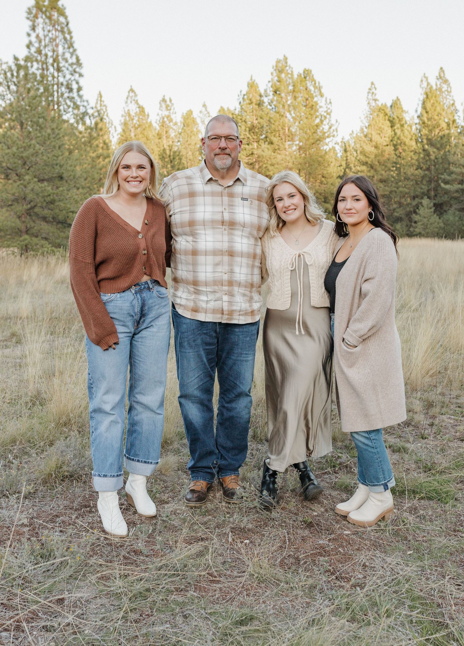 Family portrait of four people standing together in field with pine forest backdrop.