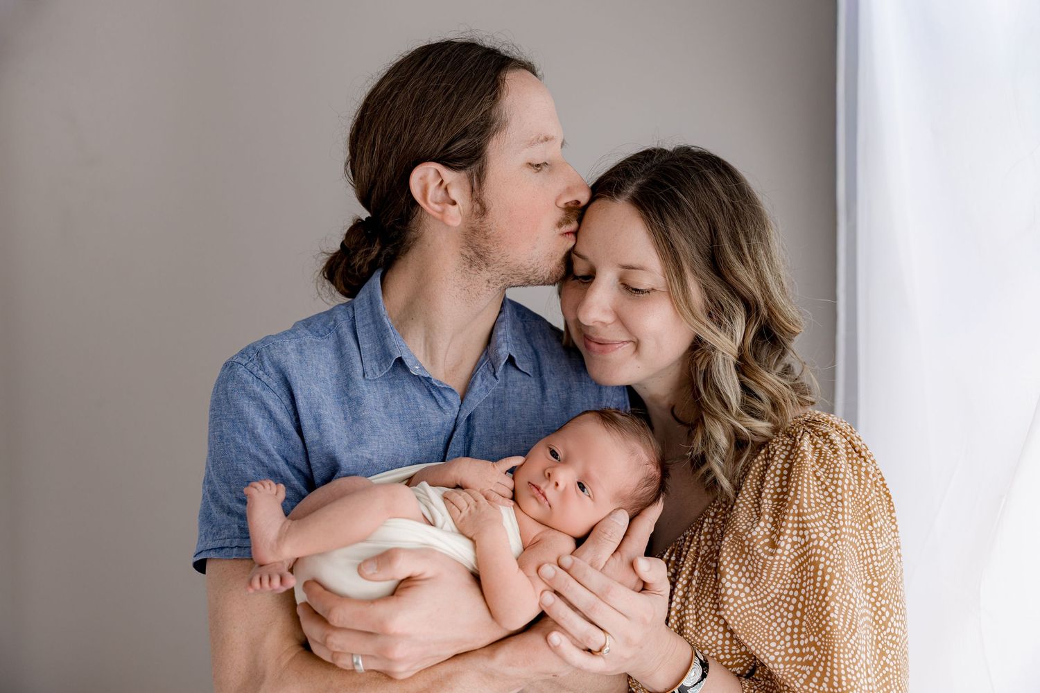 Happy couple kissing while tenderly holding their sleeping newborn baby wrapped in white blanket.