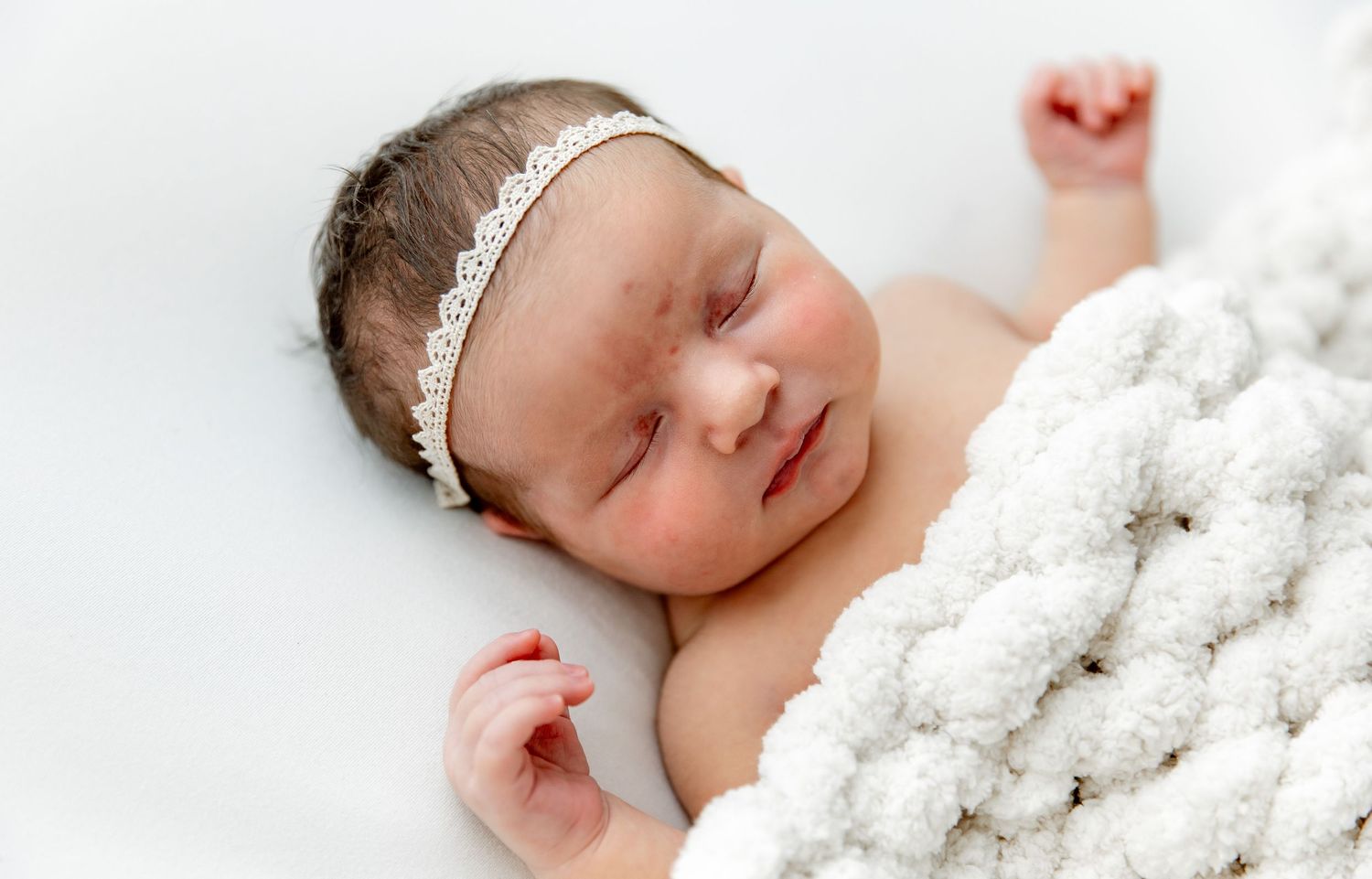 Sleeping newborn baby wrapped in soft white blanket lying on white background during professional photoshoot.
