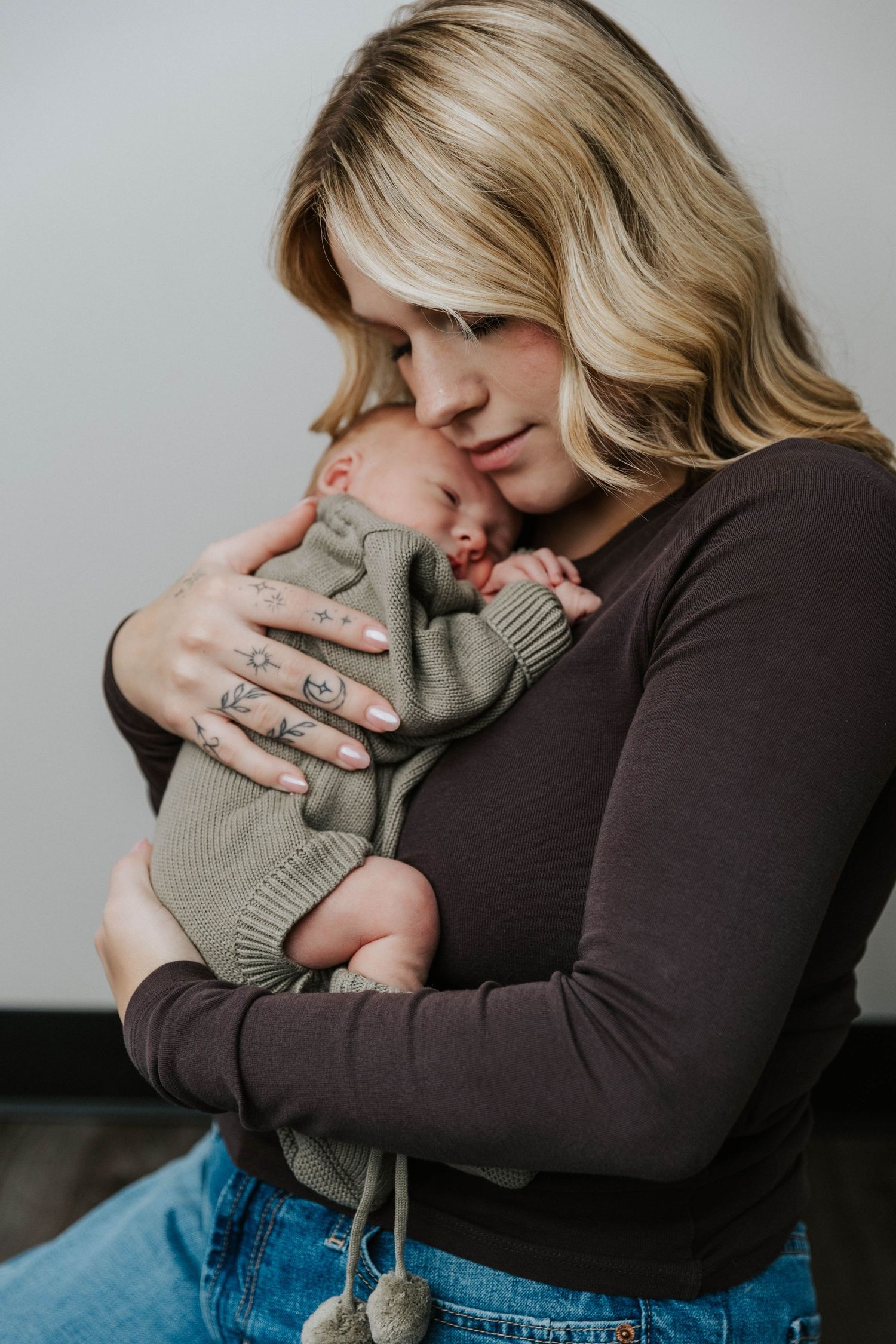 Parent wearing dark top and jeans tenderly embraces small baby in cozy gray sweater against neutral background.