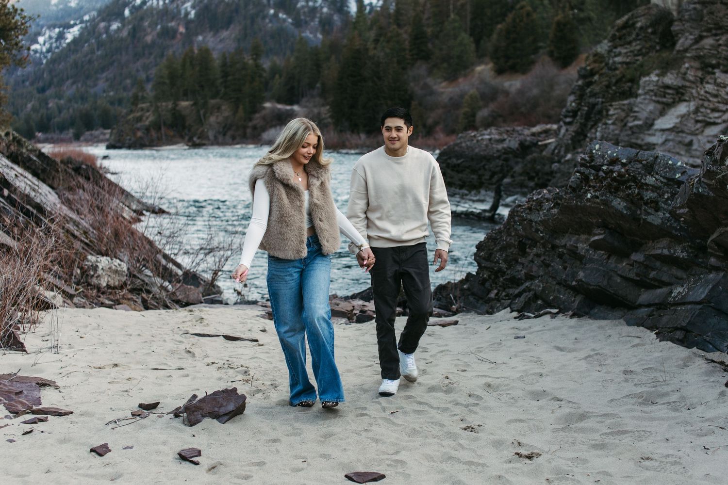 A couple walks hand in hand along a scenic mountain trail with snow-capped peaks in the background.