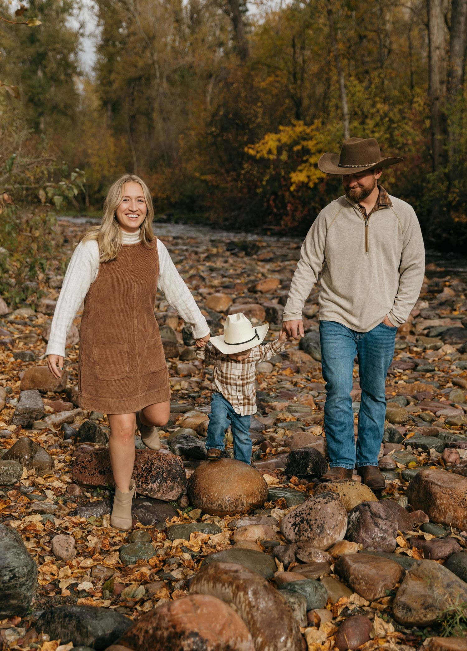 Two people explore a rocky creek bed covered in autumn leaves while wearing brown overalls and cowboy hats.