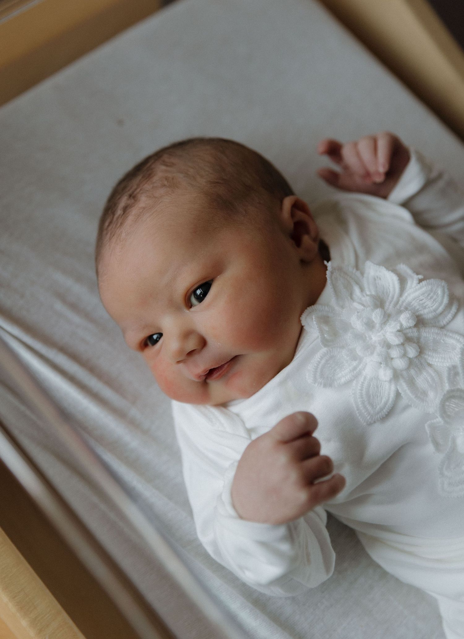 Newborn wearing white floral outfit lying on light colored bedding.