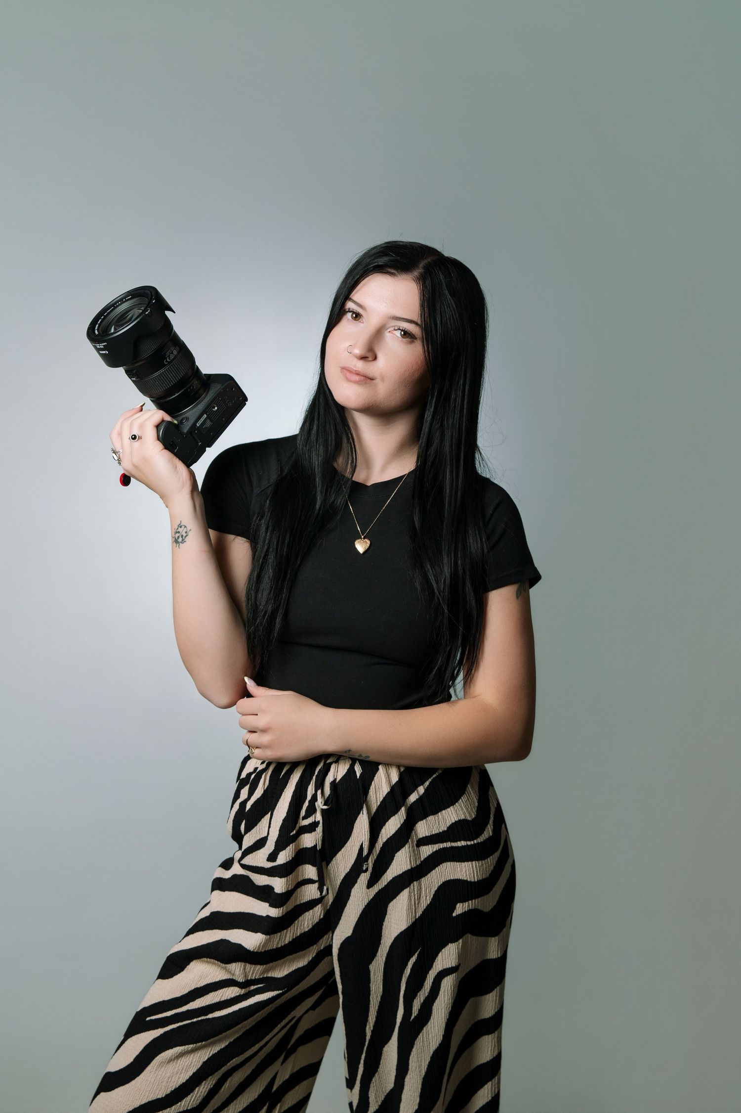 Young woman with dark hair holds a DSLR camera, wearing black top and zebra-print pants against gray background.