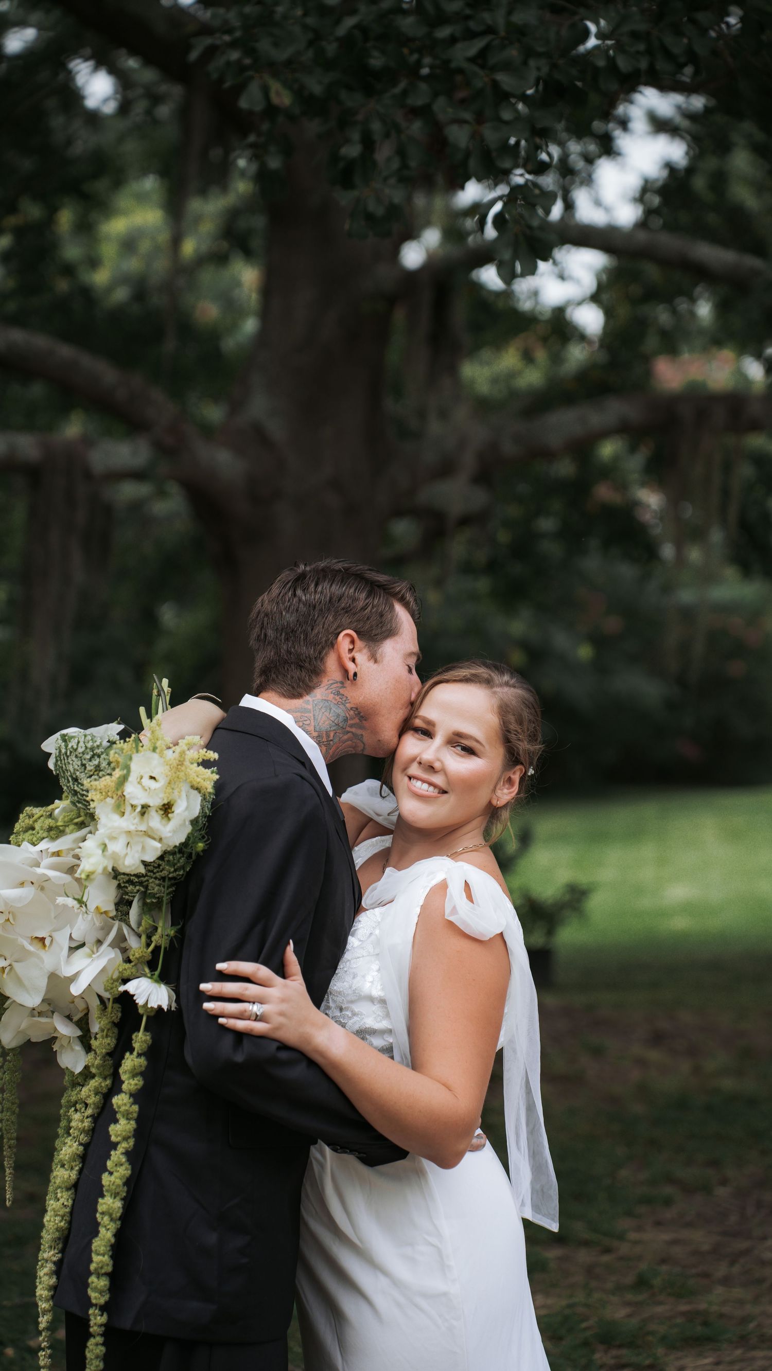 Bride smiling in white gown holding floral bouquet while groom in black suit embraces her outdoors under large trees.