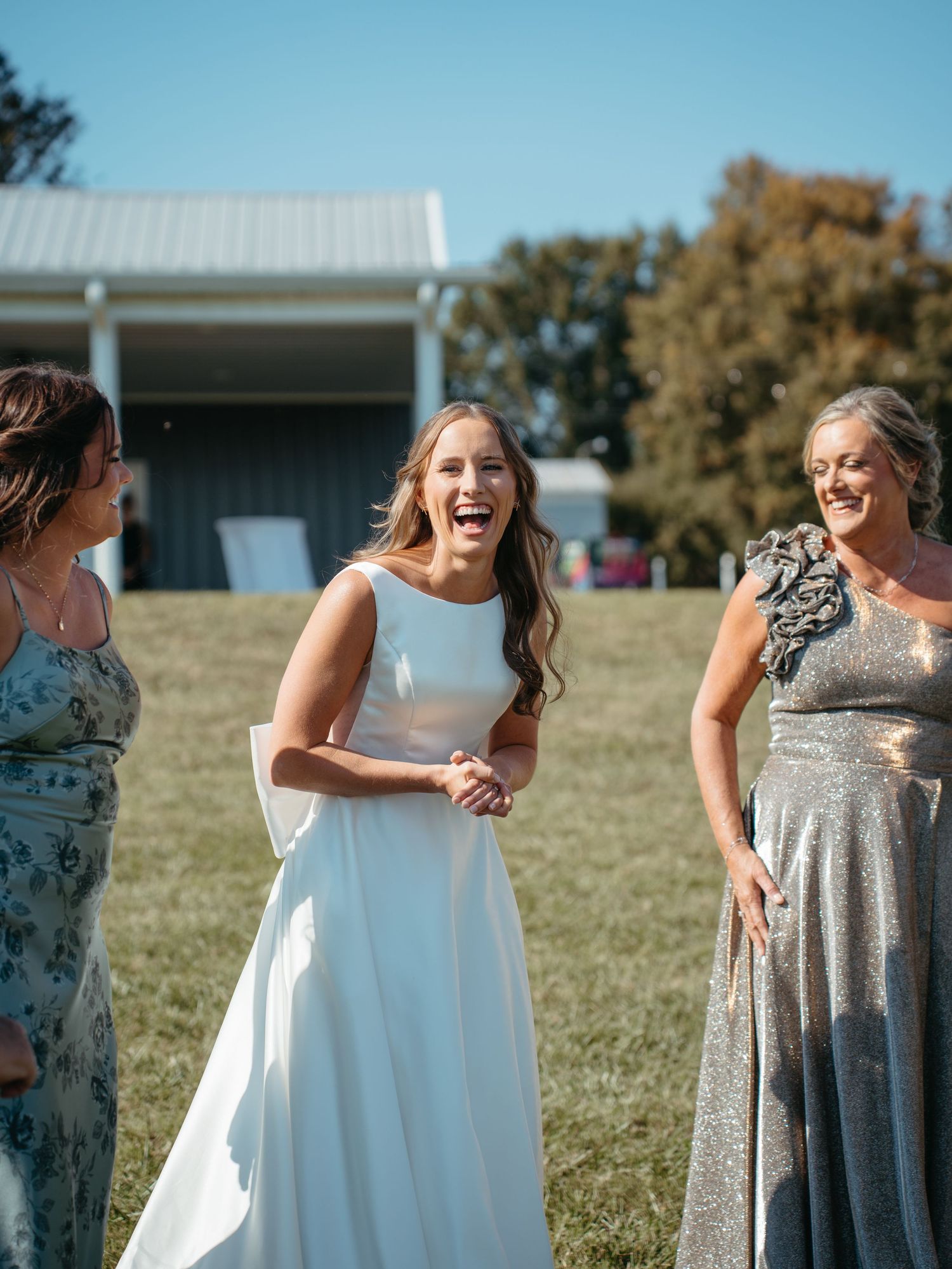 A group of bridesmaids in sparkly silver dresses and white gowns share joyful laughter in an outdoor setting.