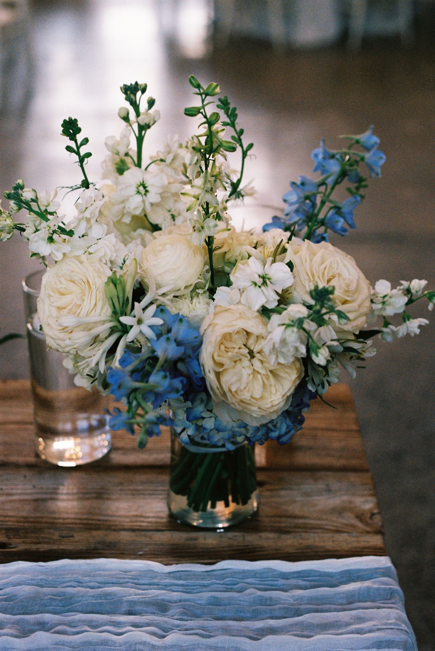 Elegant floral arrangement with white roses and blue delphinium flowers in a glass vase on wooden surface.