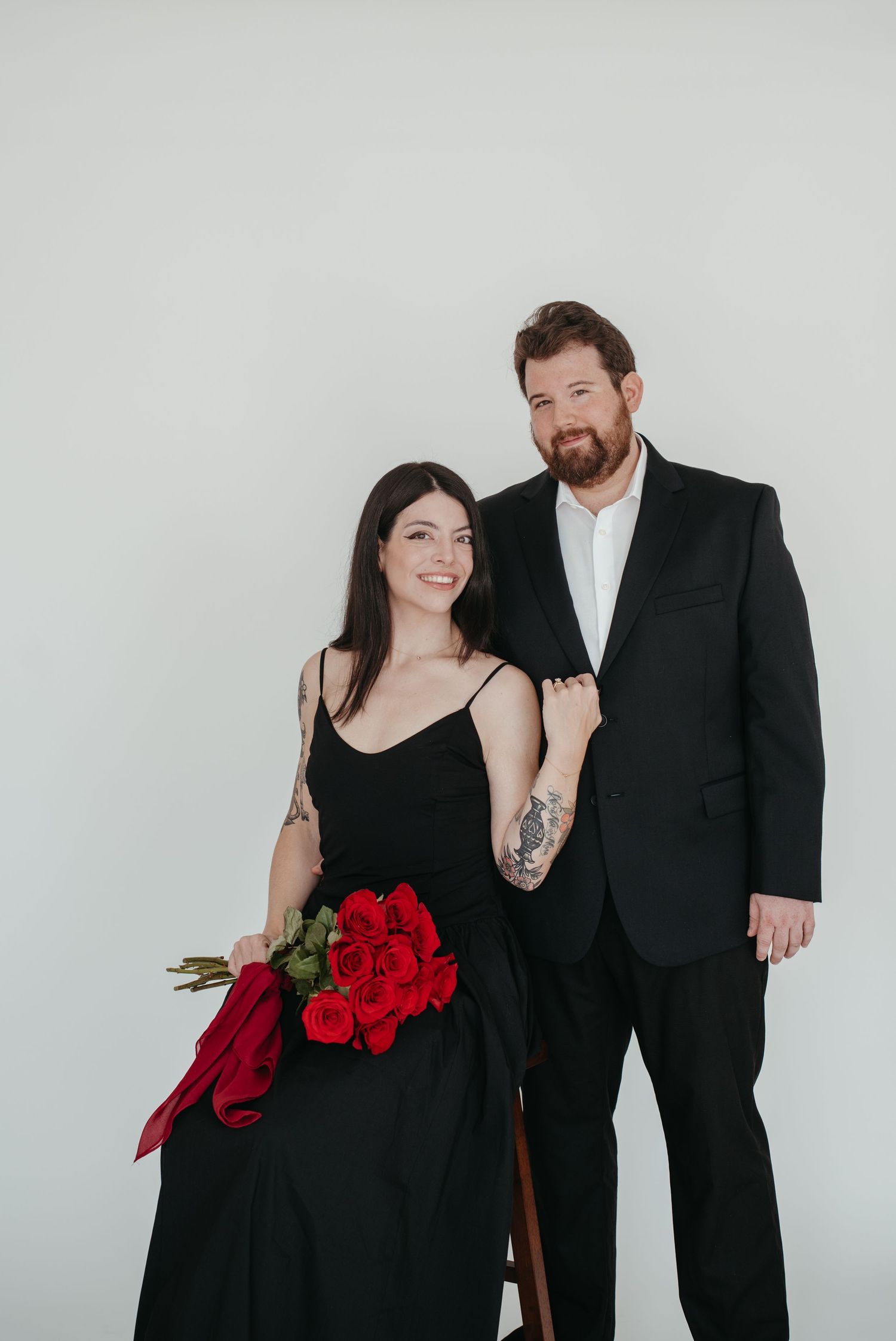 Formal couple in black attire poses with red roses against white backdrop.