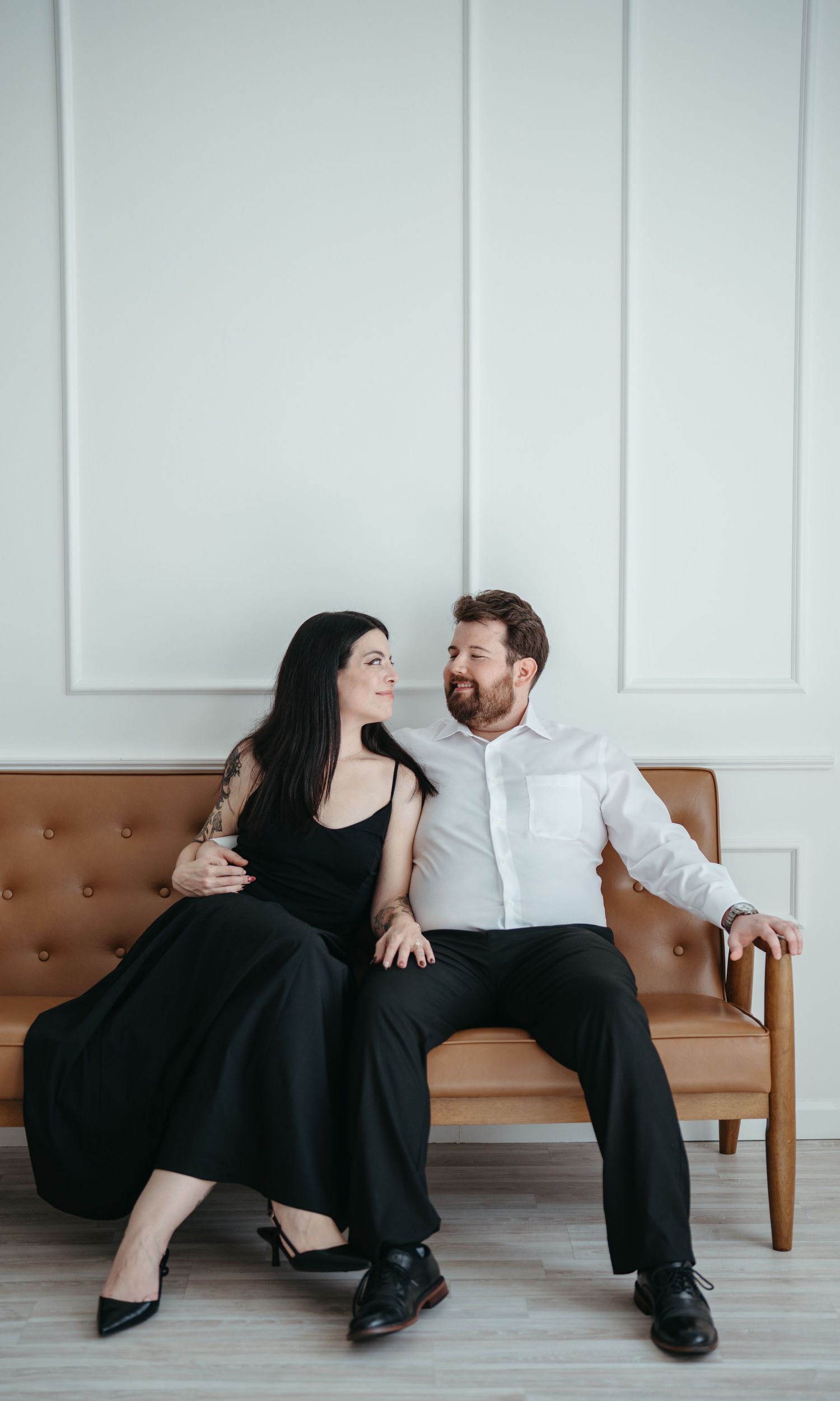 A couple in formal black and white attire shares intimate moments while sitting on a brown leather bench.