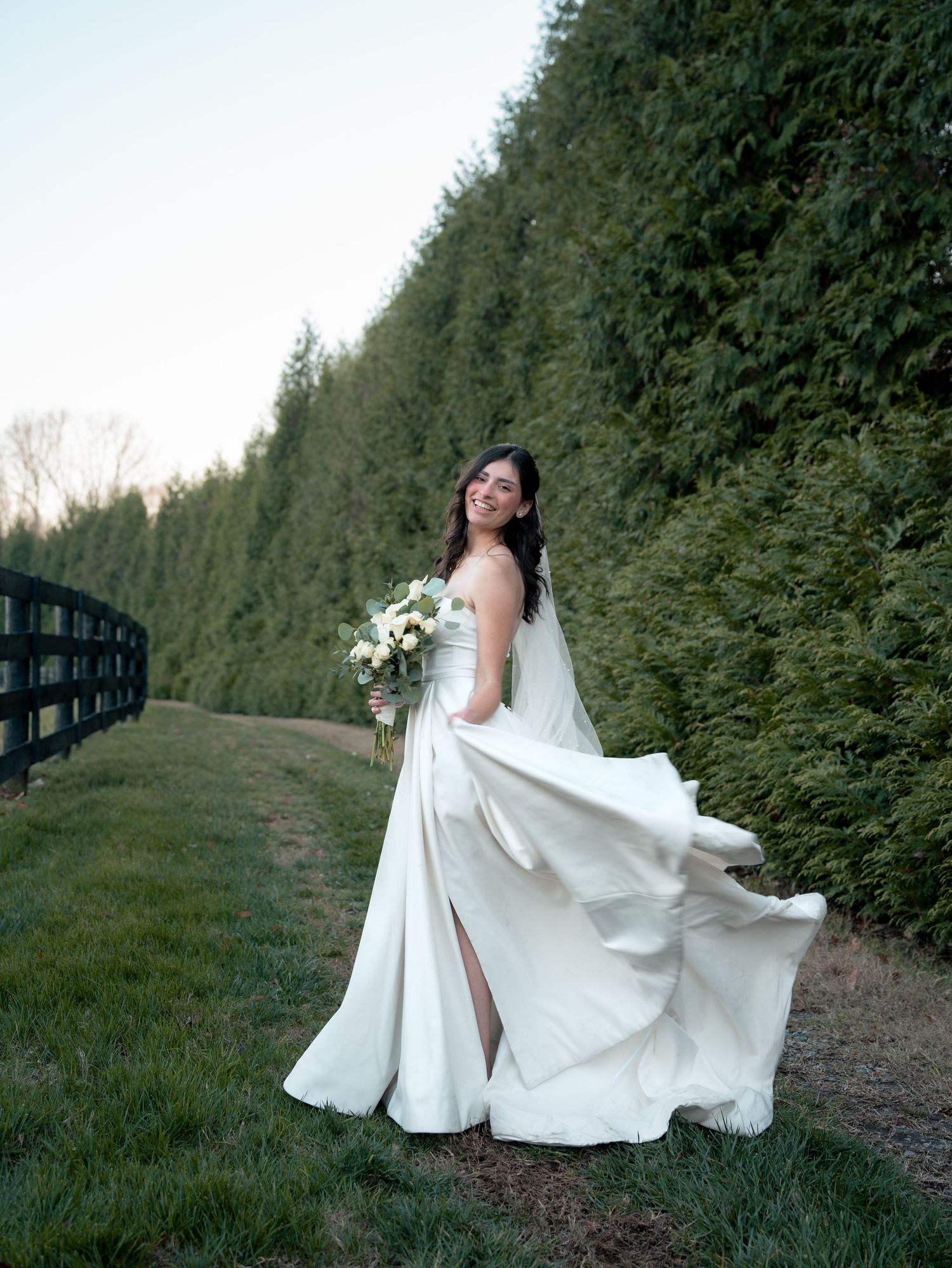 A flowing white wedding dress billows gracefully against a backdrop of tall evergreen hedges in a garden setting.