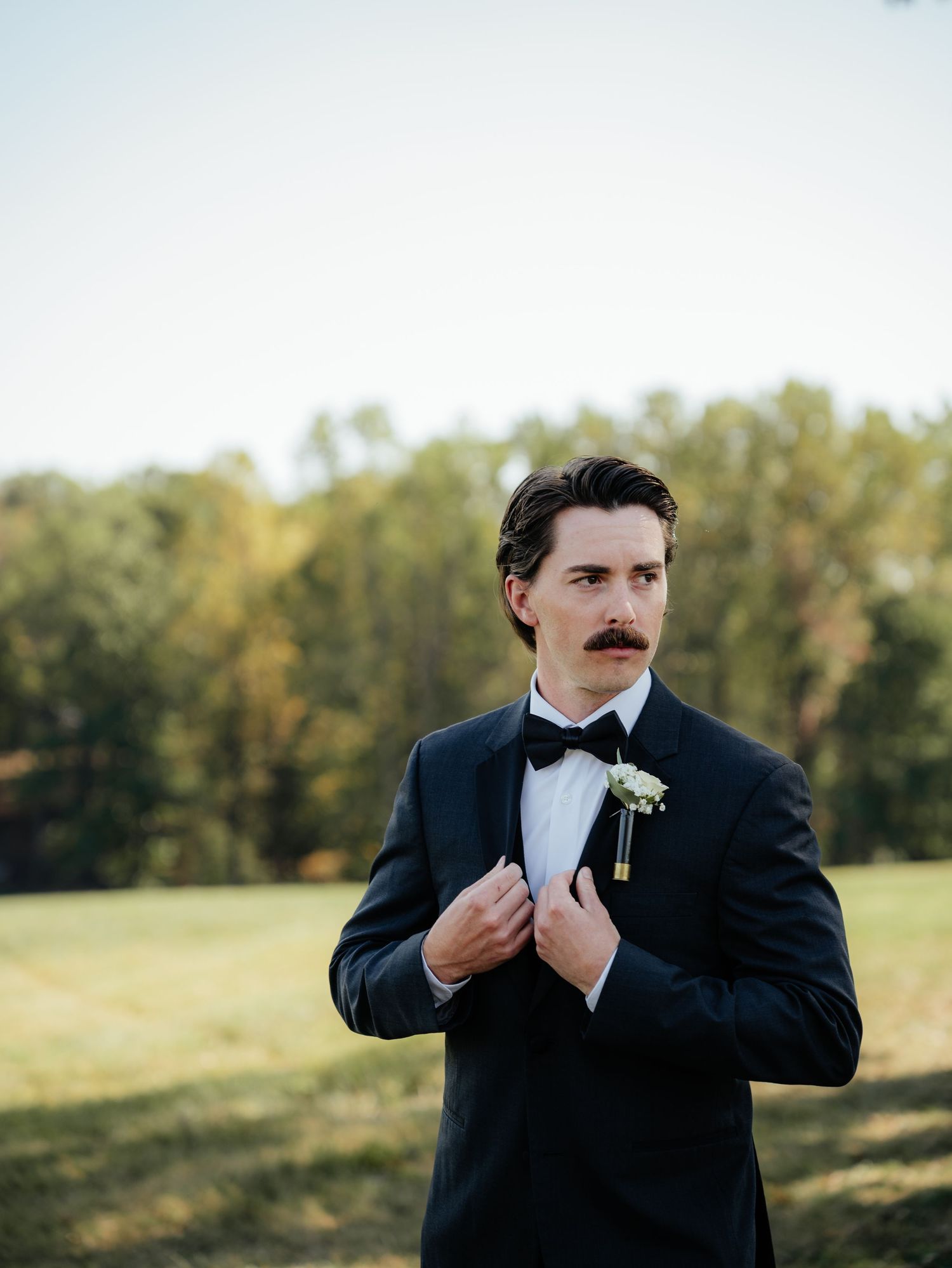 Dapper groom in black tuxedo with bow tie and boutonniere adjusting jacket outdoors on wedding day.