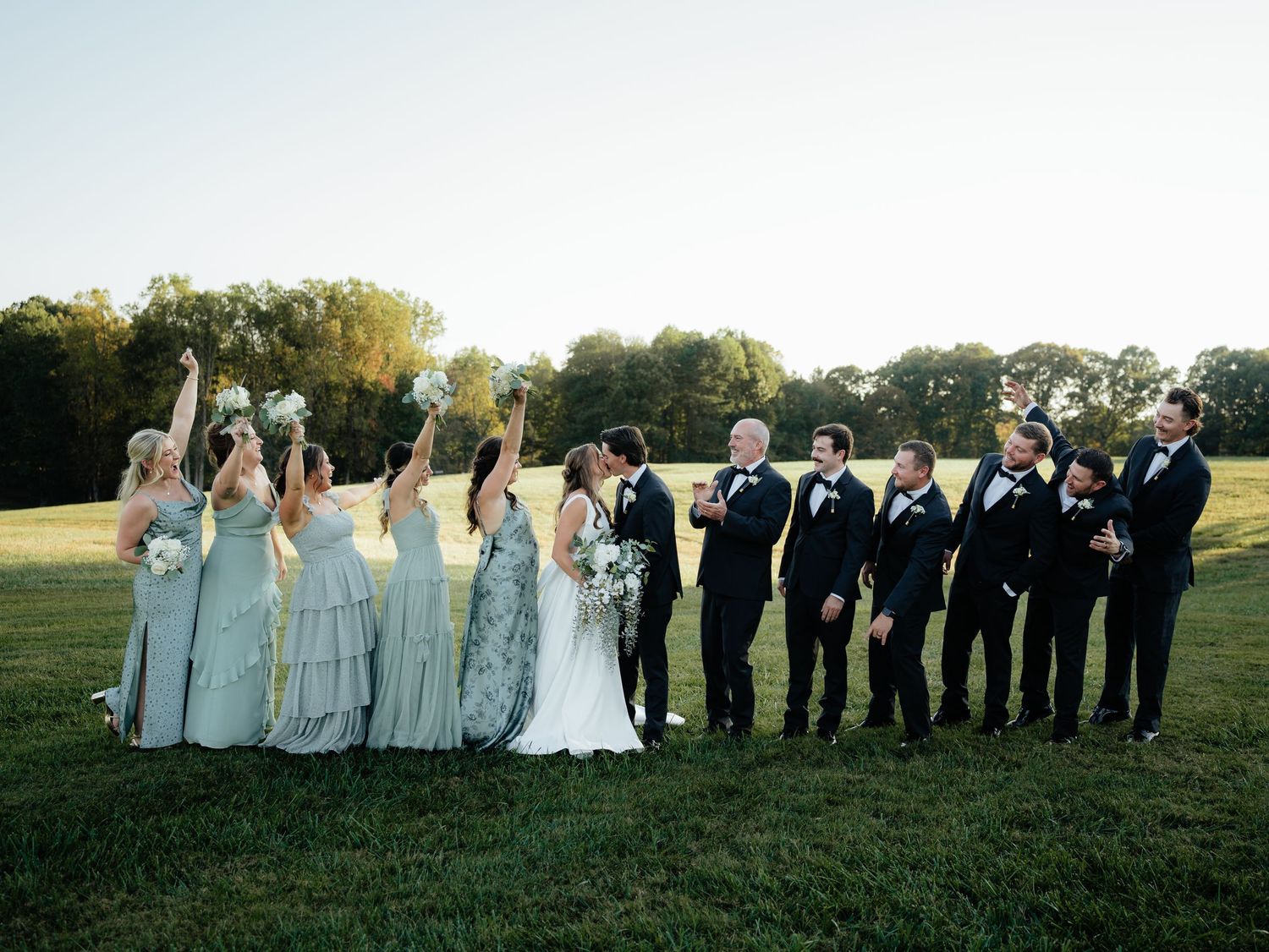 Wedding party poses outdoors on green lawn, bridesmaids in sage gowns cheering beside bride and groomsmen in black tuxedos.