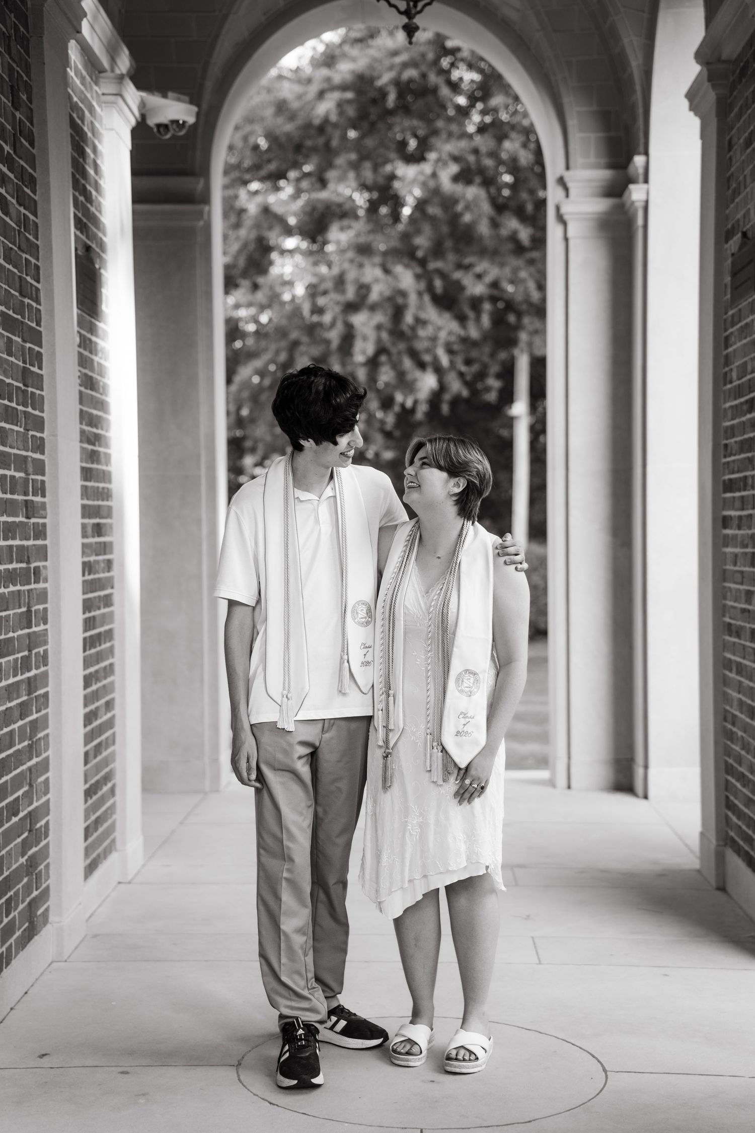 Black and white photo of two graduates in stoles standing together under an arched brick colonnade.