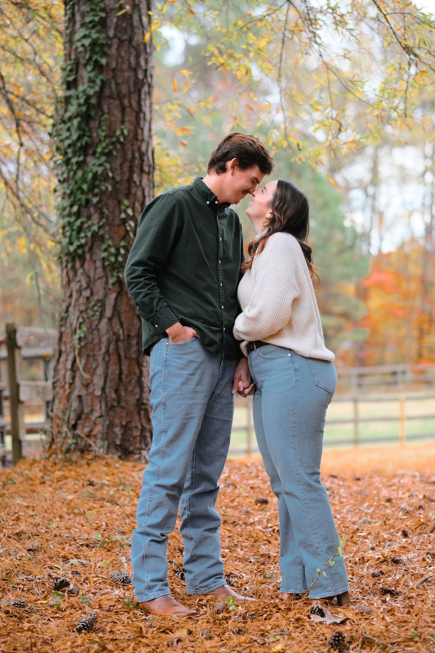 Couple in casual attire sharing intimate moments while standing on fallen leaves in a forest setting.