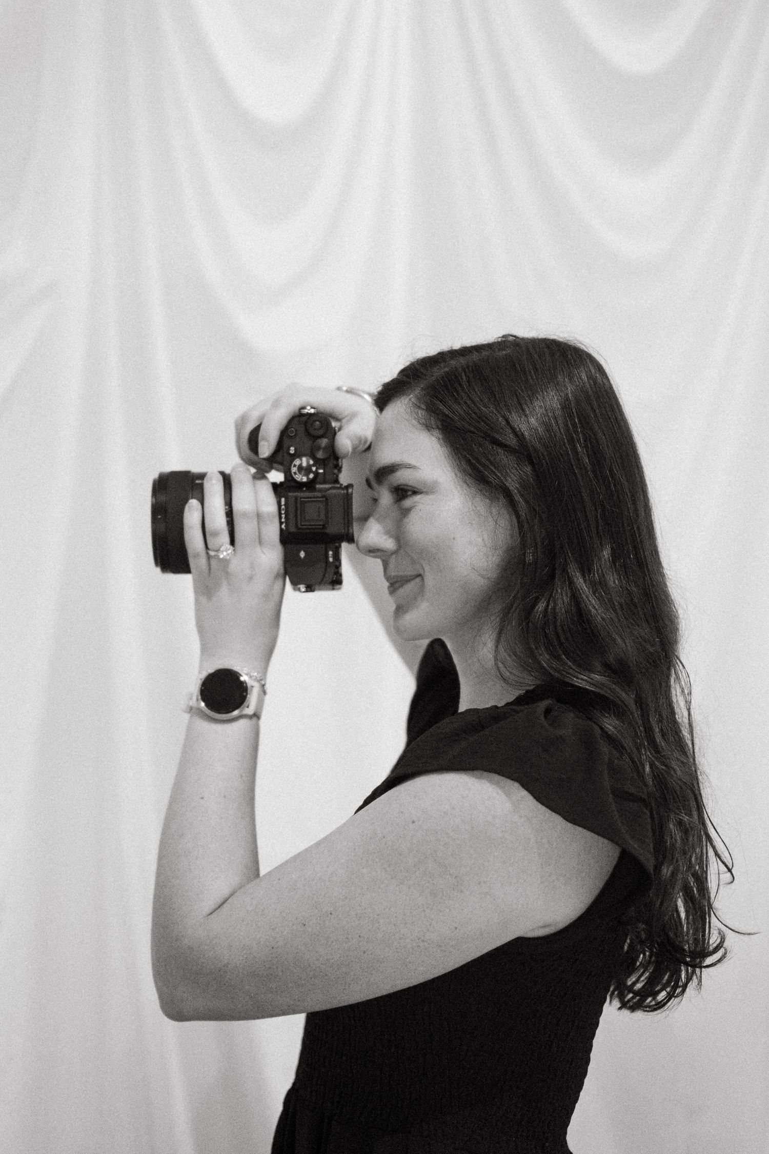 A photographer in a black sleeveless top looks through a camera lens against a white background in a profile view.
