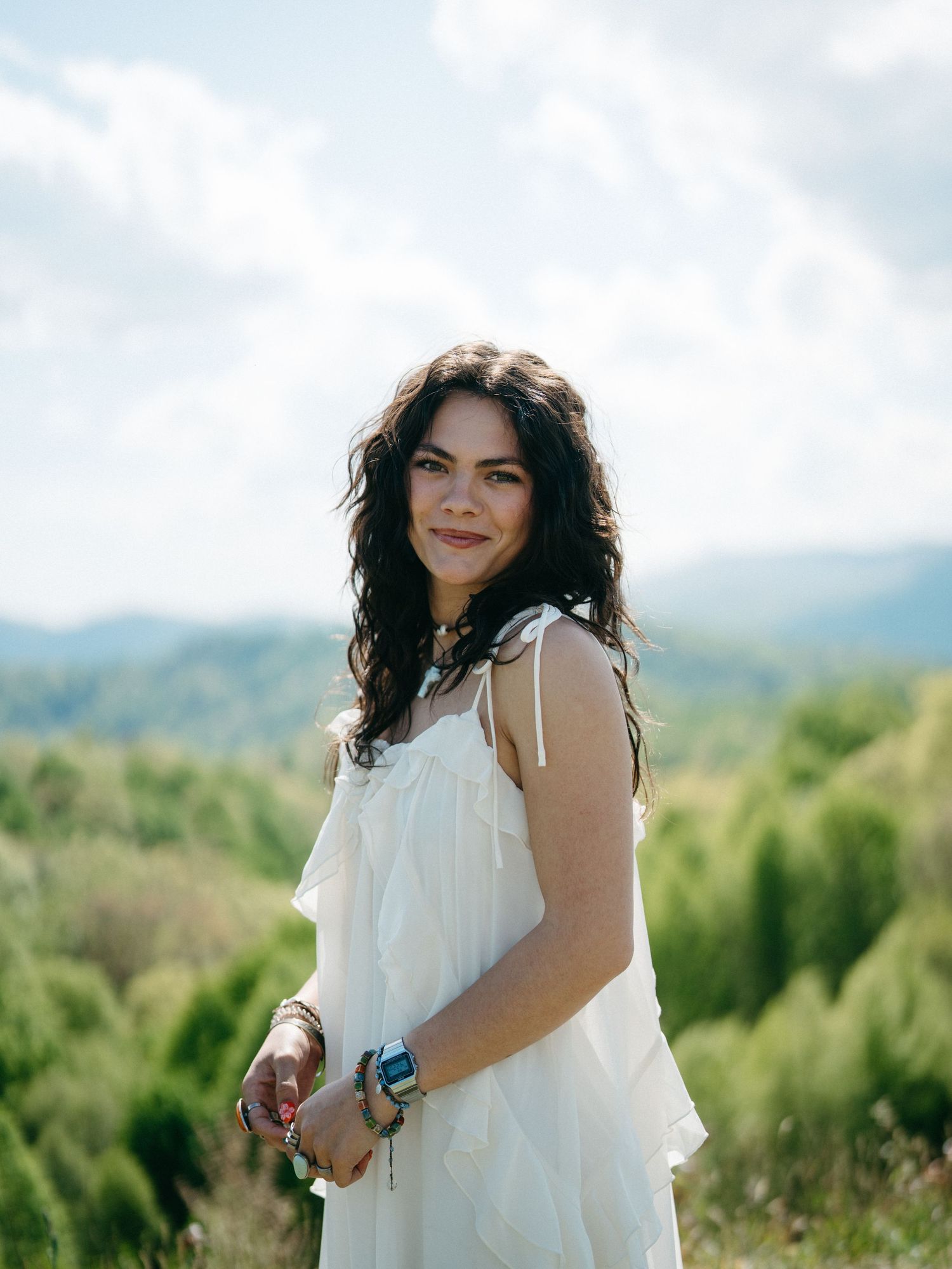 Young woman in white dress smiling outdoors with lush green hills and cloudy sky in background.