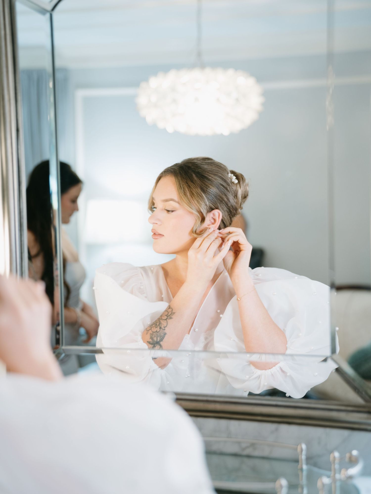 Bride in white robe putting on earrings while looking in a mirror, soft elegant bridal getting ready moment.