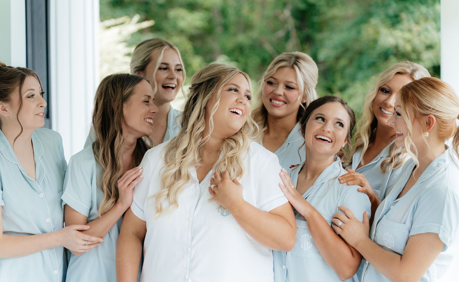 A group of women in light blue and white dresses share a joyful moment together while getting ready.