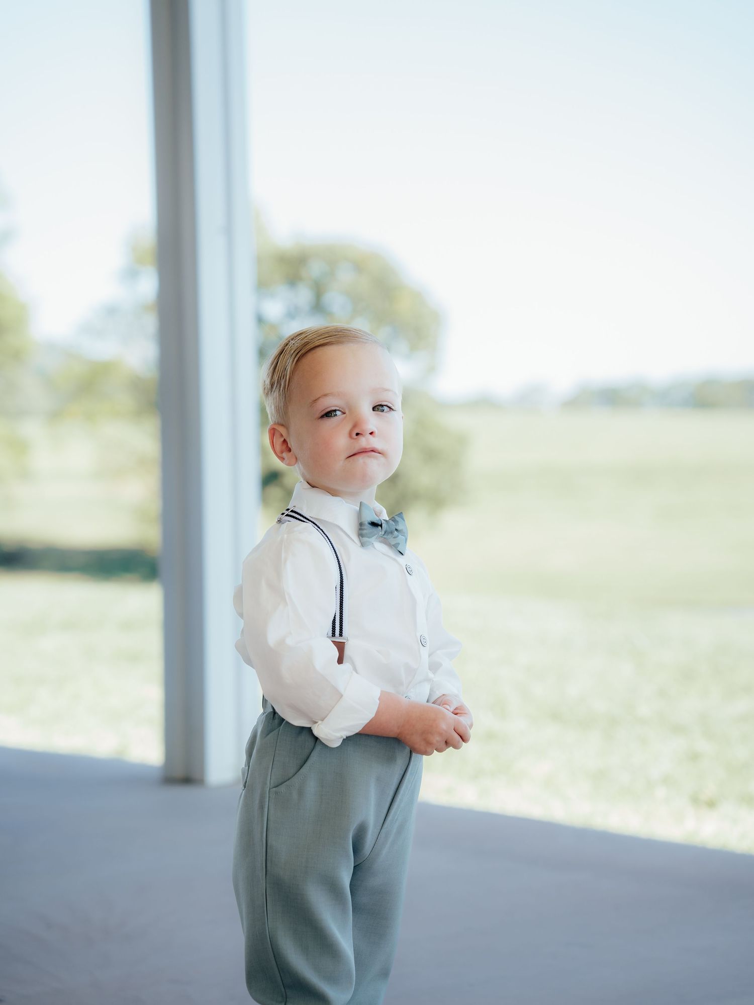 A small child wearing a gray vest and white shirt stands in front of a bright window.