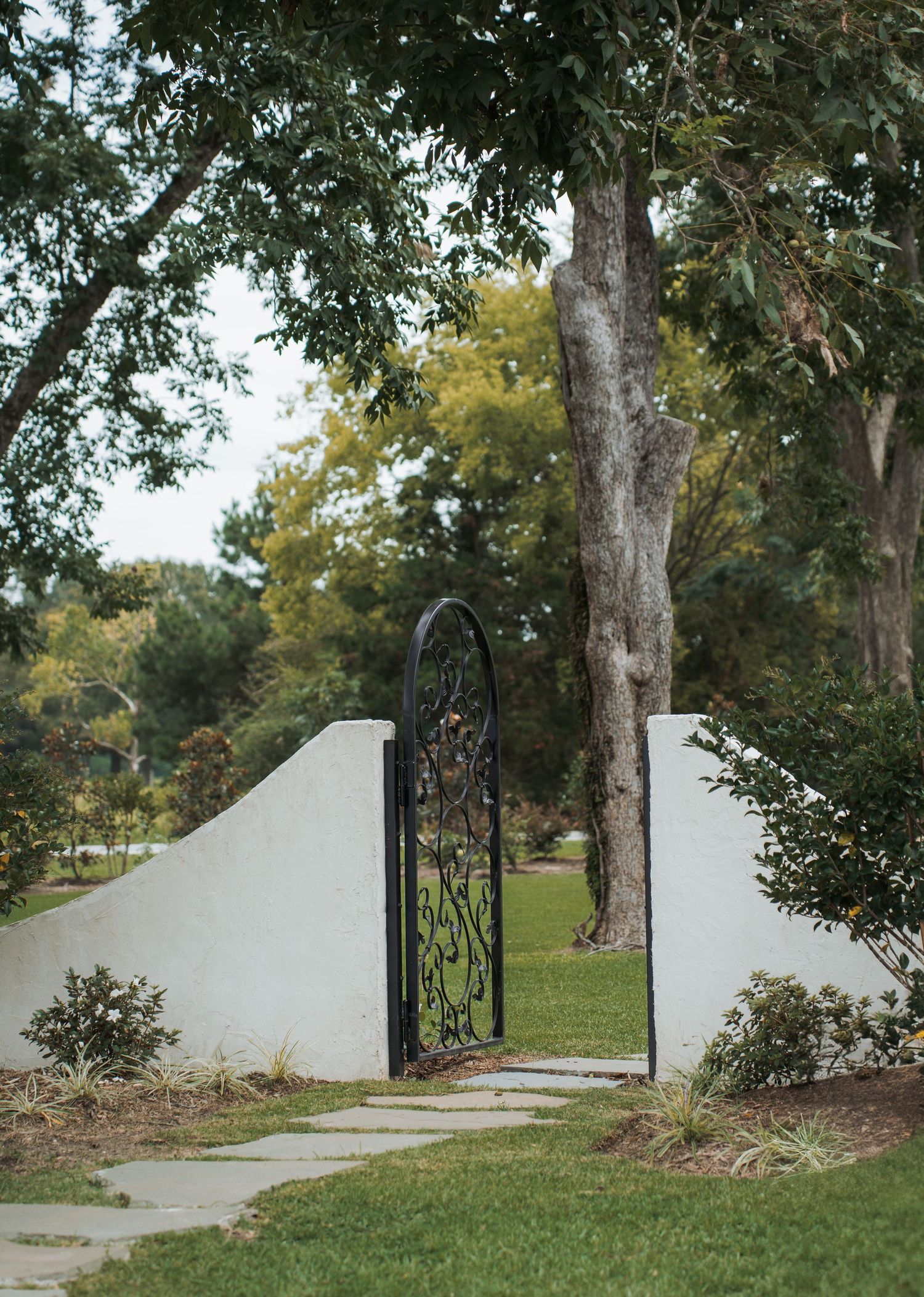 A curved white stucco wall with ornate black iron gates leads to a garden path surrounded by mature trees and landscaping.