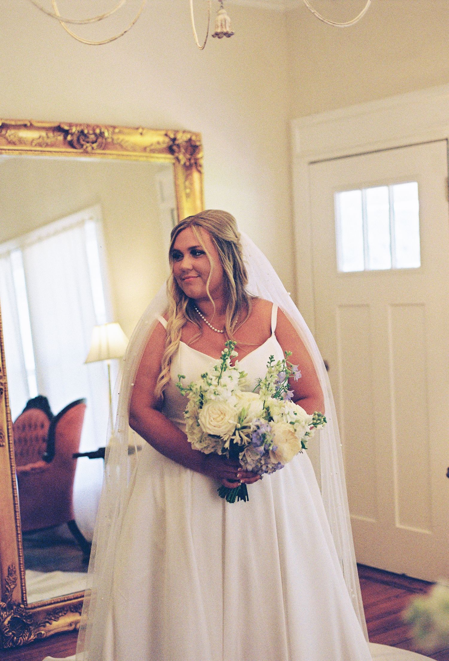Bride holds a white and green bouquet in a sunlit room with cream walls.