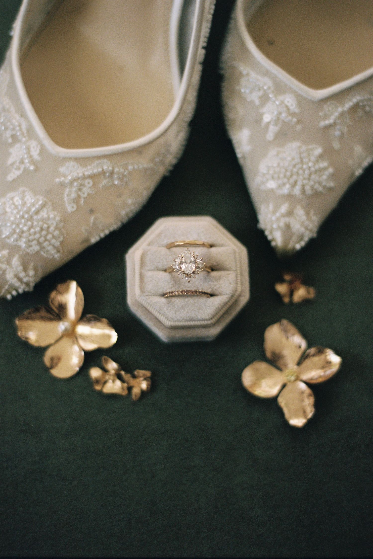 Elegant white lace bridal shoes with delicate beading details arranged in a row with gold leaves on dark background.