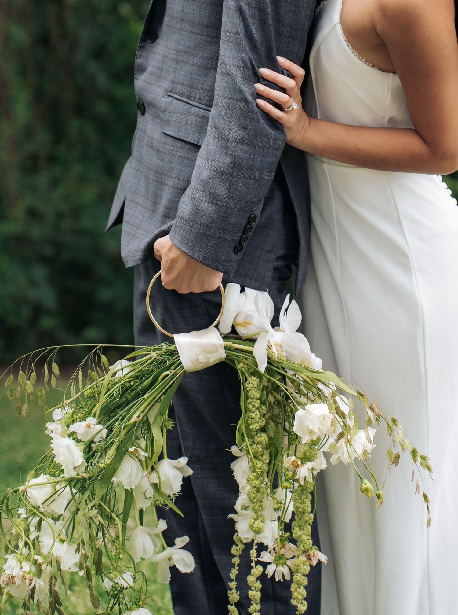 A couple in wedding attire stands close together while holding a cascading white floral bouquet with greenery.