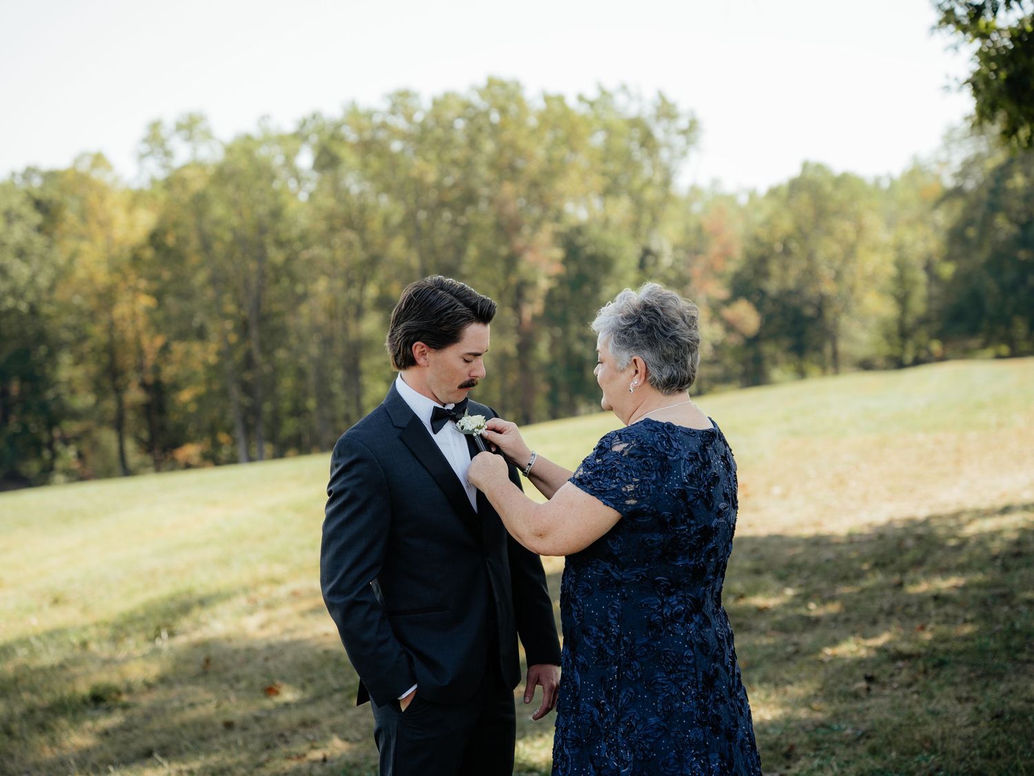 A person in a black suit getting their boutonniere pinned on by someone wearing a navy blue dress in an outdoor setting.