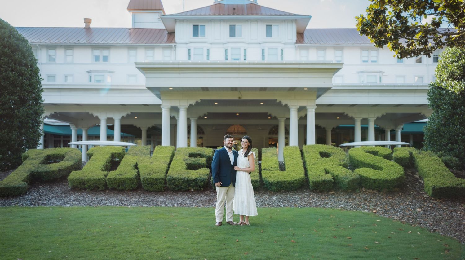 Couple embracing in front of Pinehurst Resort topiary sign on lush green lawn with historic white clubhouse backdrop.