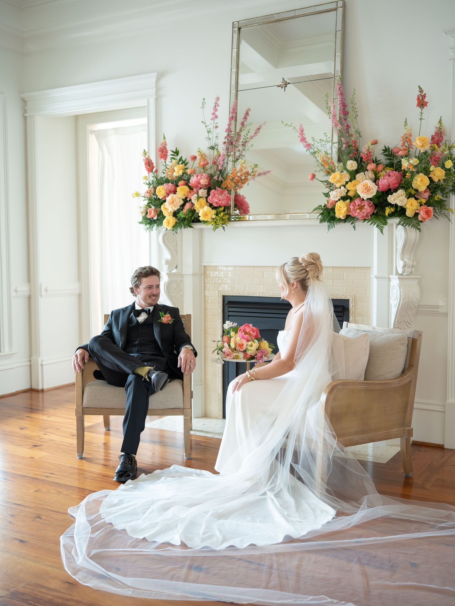 Bride and groom seated by a white fireplace adorned with colorful floral arrangements, bride in elegant gown with veil.