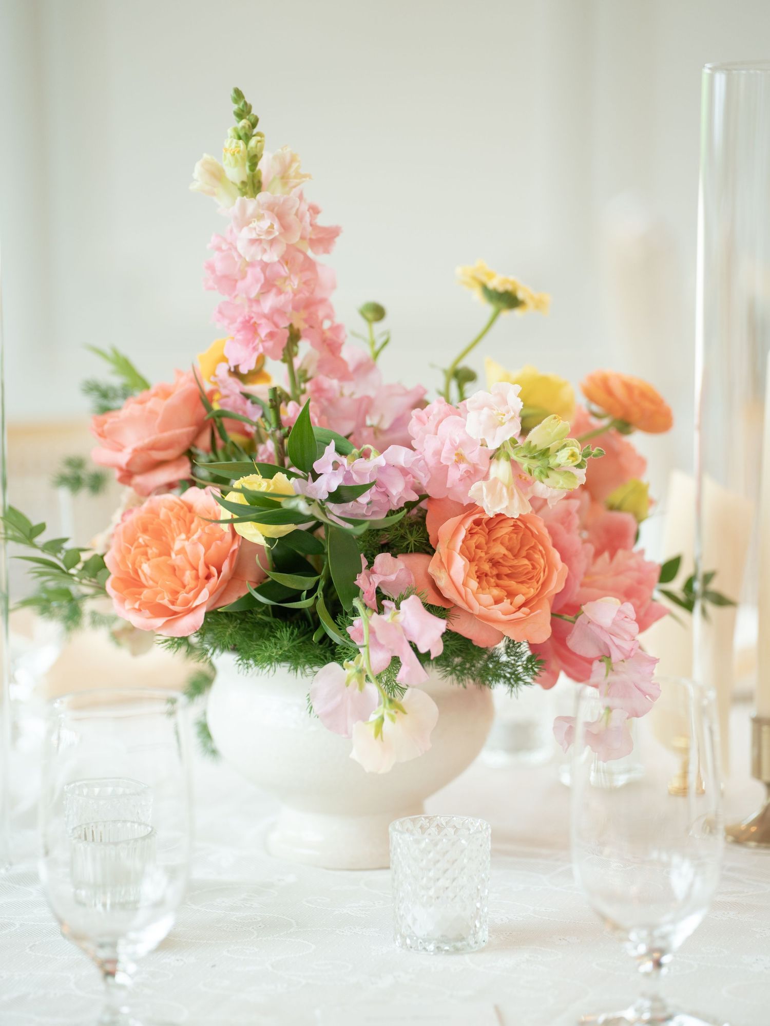 Elegant floral centerpiece with peach garden roses, pink snapdragons, and ranunculus in a white bowl on a reception table.