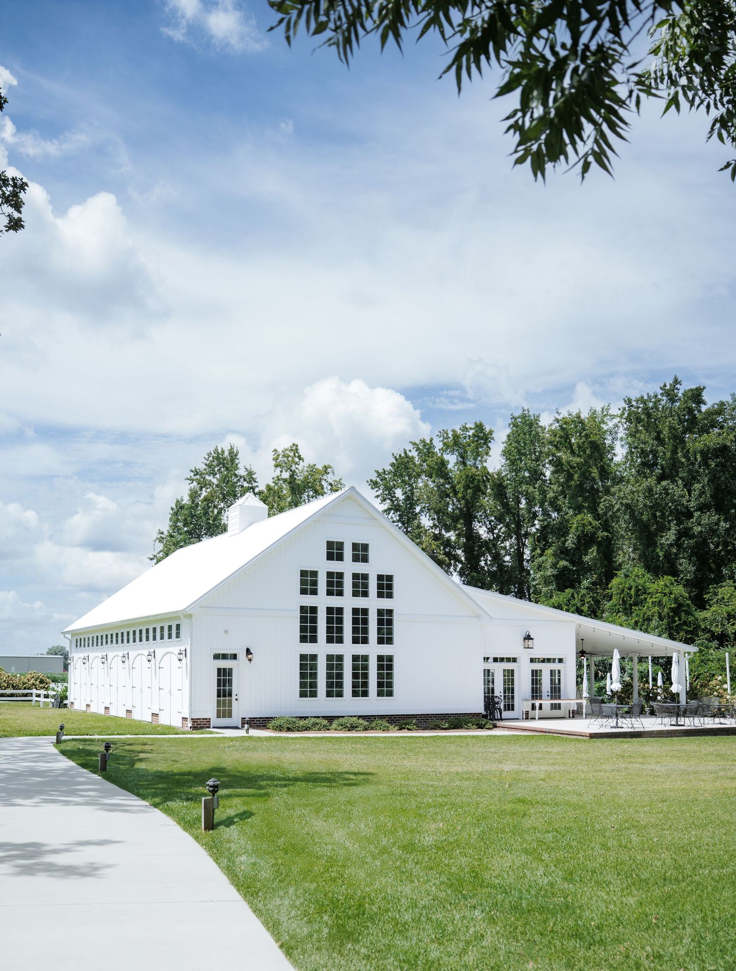 Modern white barn-style event venue buildings with black windows set on a lush green lawn under blue skies.