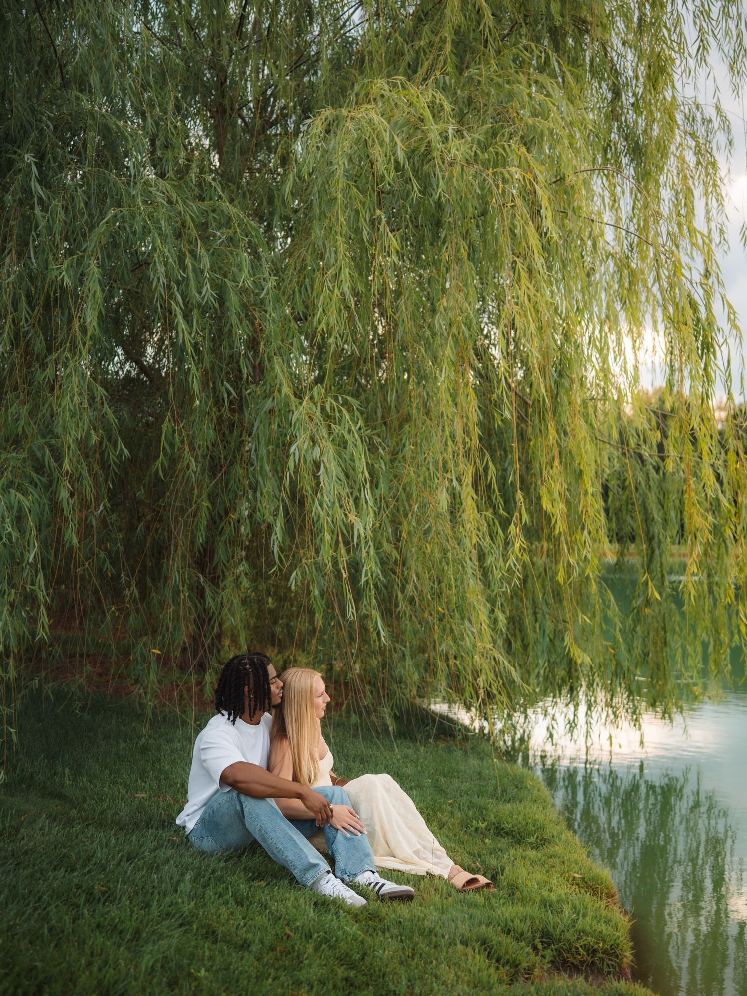 Couple sitting together under a weeping willow tree by a serene lake at golden hour.