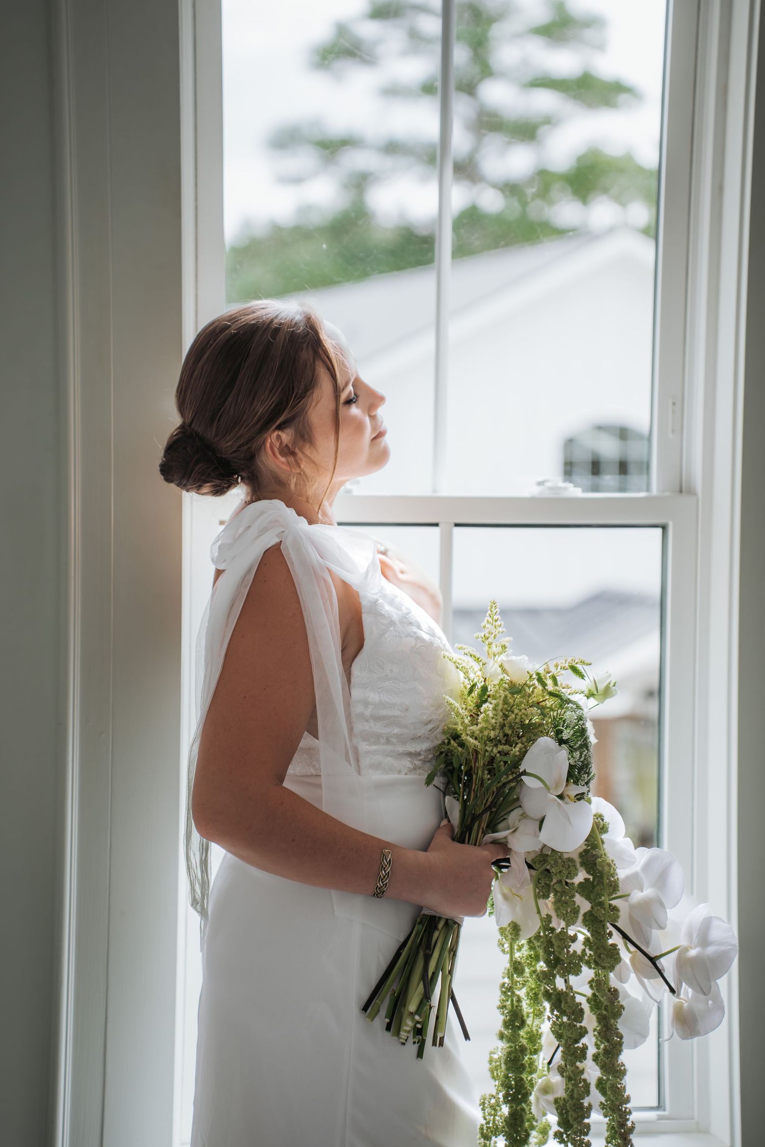 Elegant figure in white dress holds bouquet while posing by large windows.