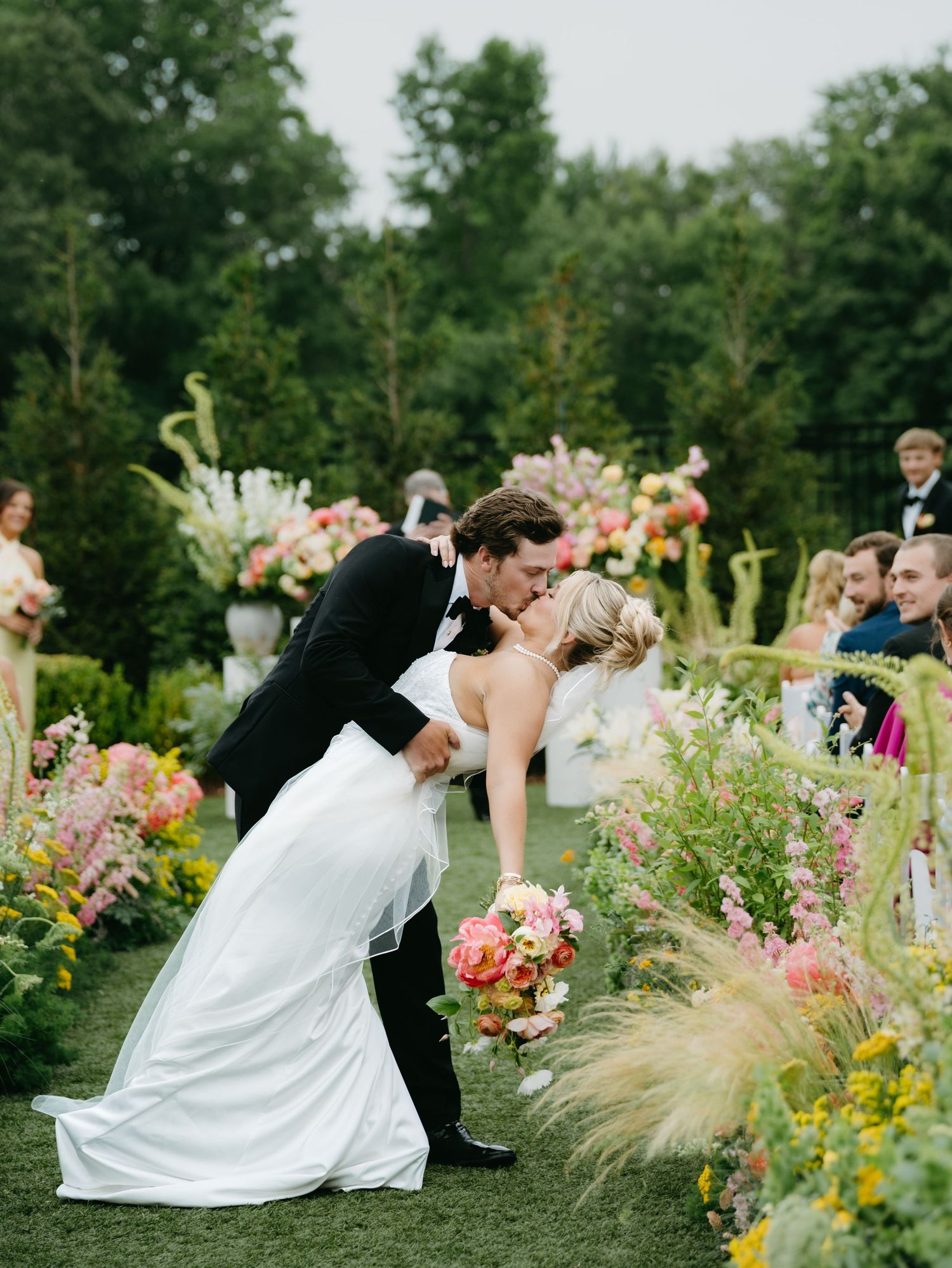 Bride and groom share a romantic dip kiss at outdoor garden wedding ceremony surrounded by colorful floral arrangements.