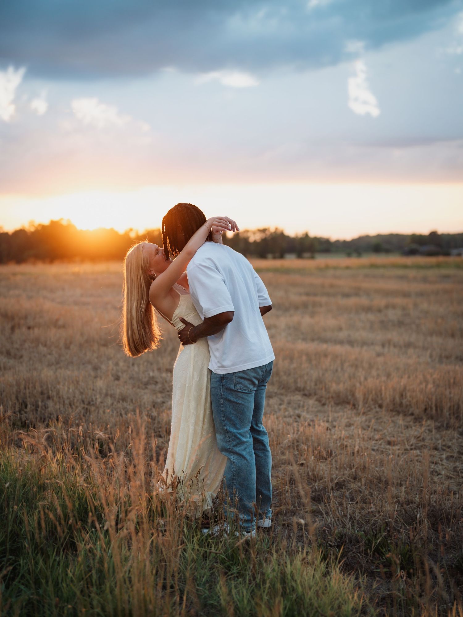 Couple dancing and kissing in a golden field at sunset, woman in white dress, man in jeans and white tee.