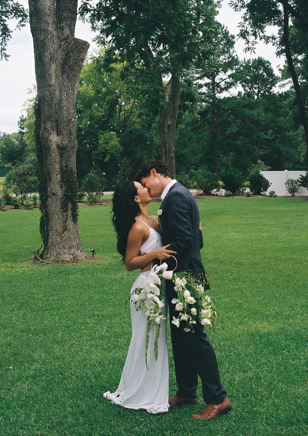 A romantic wedding couple shares an intimate moment on a grassy lawn beneath tall trees.