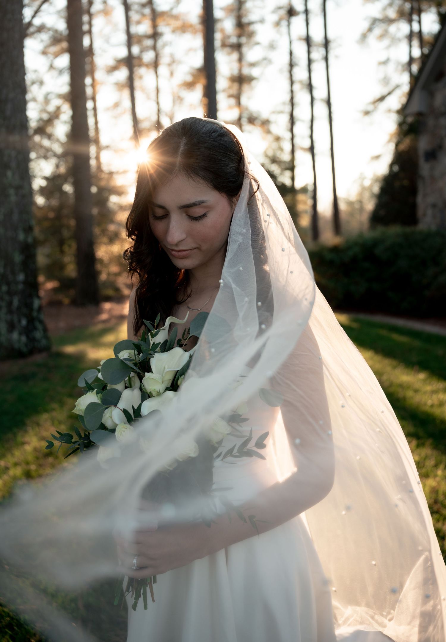 A bride in a flowing white veil holds a bouquet of white flowers against a sunlit forest backdrop at golden hour.