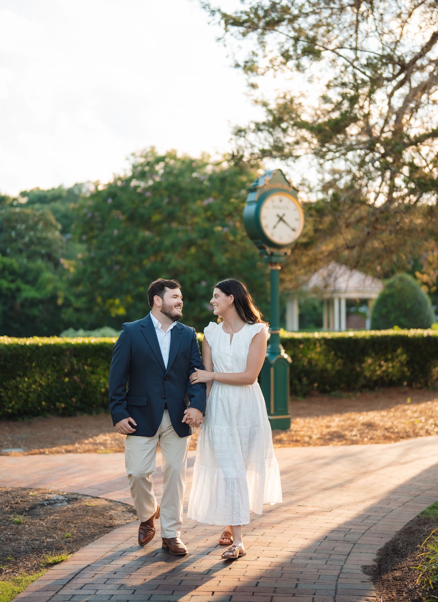 Couple holding hands walking on brick path with green clock tower and lush trees in background during golden hour.